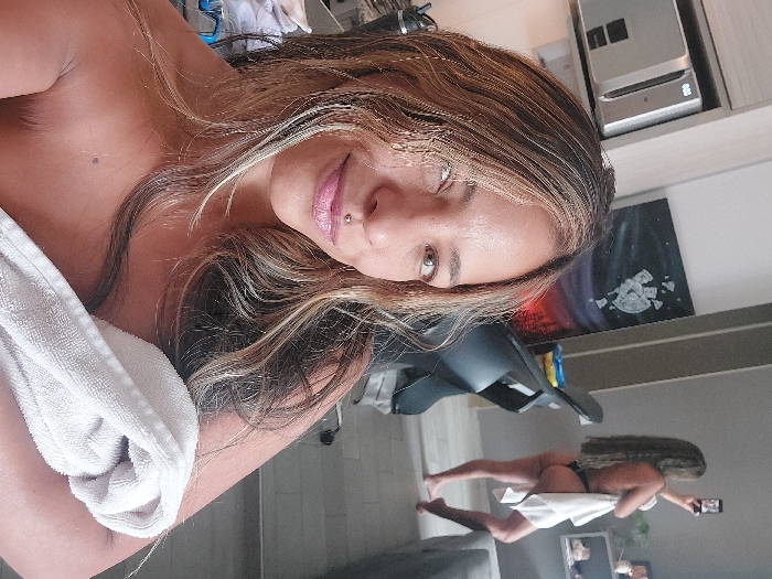 A selfie shows a woman with highlighted brown hair, a pierced nose, and pink lipstick, partially covered by a white towel. In the background, a mirror reflects the same woman in a full body shot, holding a phone taking a picture. She is wearing black underwear and also covered by a white towel. A black office chair is visible in the mirror's reflection. Above the woman is some sort of printer or filing cabinet. A canvas with space art is mounted on the wall.