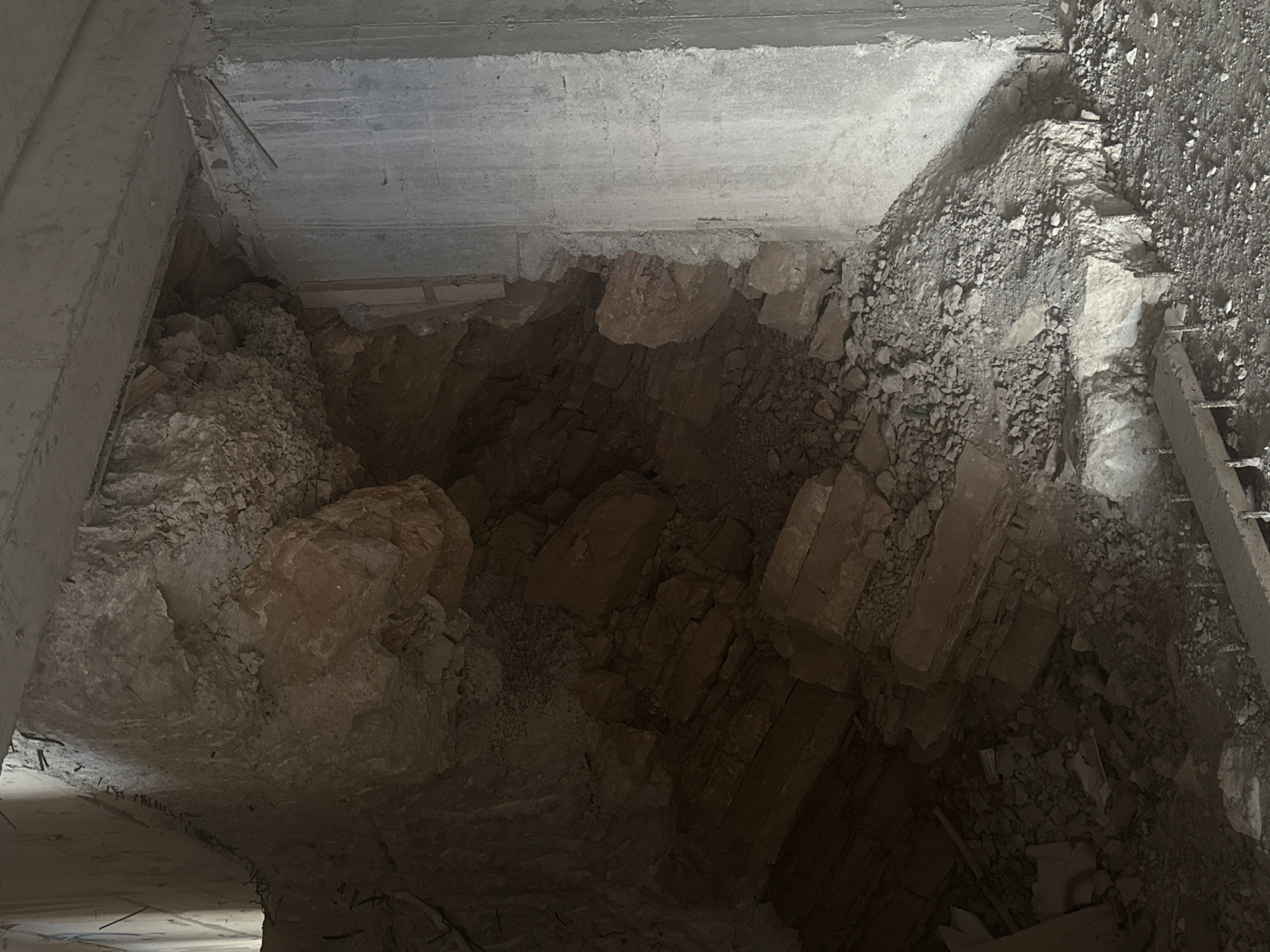 This image shows a collapsed section of a concrete structure, possibly part of a building or tunnel, in Petrovac na Moru, Montenegro. The collapse has exposed layered rock formations and debris. The scene is dark and appears to be an interior or underground space. There are no people visible. The environment is dusty and filled with rubble, indicating recent structural failure. The concrete elements appear aged and show signs of wear and tear, with exposed rebar visible in some areas. The overall impression is one of disrepair and destruction.
