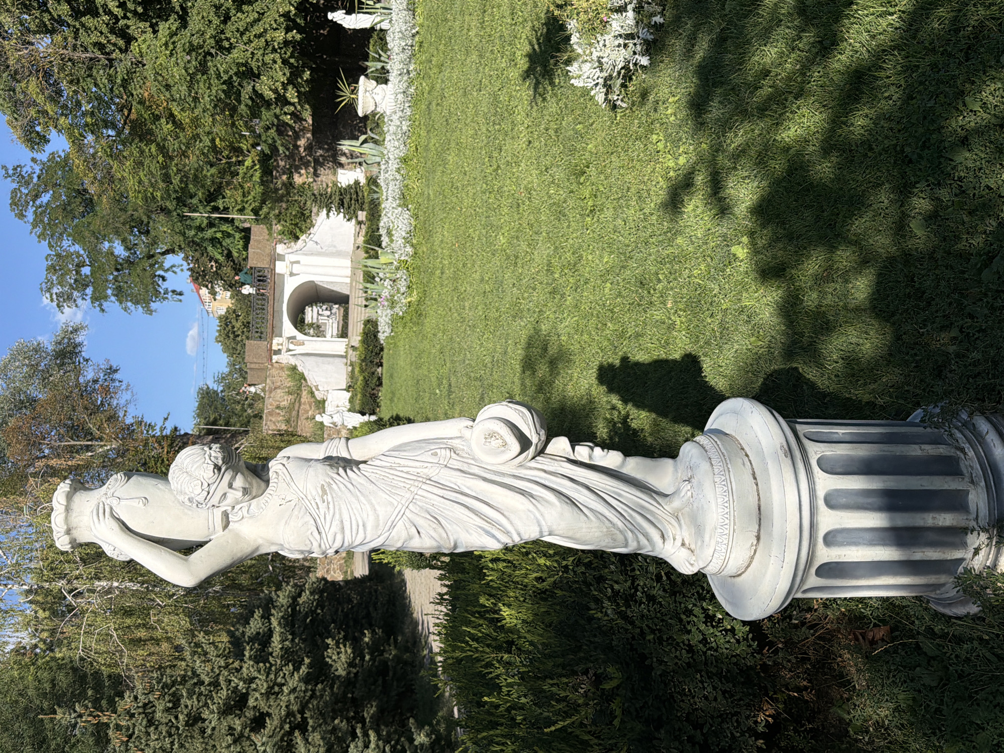 The image shows a statue of a woman holding a vase on her head, in the Botanical Garden of Rostov-on-Don, Russia. The statue is made of white stone and stands on a pedestal. The woman is wearing a draped toga and has her hair tied up in a bun. She is holding a round object in her left hand. The statue is in a garden with green grass and trees. In the background, there is a white arched bridge with people walking on it. The sky is blue and clear. The overall impression is one of a peaceful and beautiful day in a park.