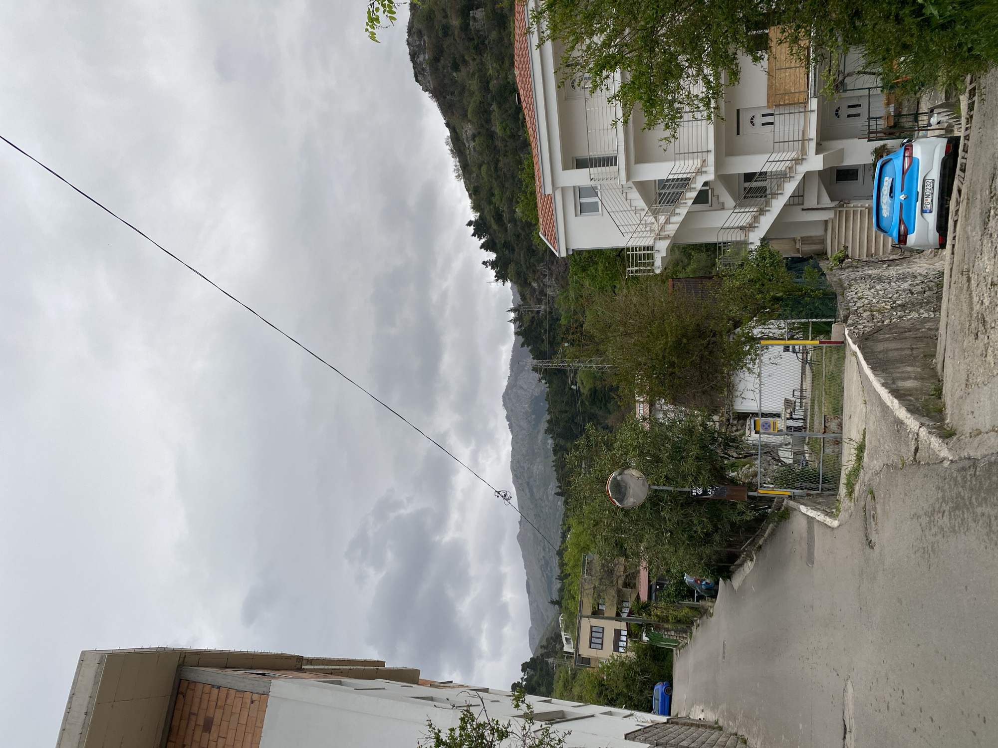 The image shows a street in Budva, Montenegro, under an overcast sky. In the background, a mountain range is partially obscured by clouds. A power line stretches across the upper portion of the frame. On the right side of the street is a multi-story white building with red roof tiles and a metal staircase. Parked adjacent to the building is a blue and white Renault car with the license plate PG-ND232. Further down the street, a convex mirror is mounted on a pole, providing visibility around a blind corner. A gate and a chain-link fence are also visible. On the left side of the street, a building under construction with exposed brickwork is partially visible. The asphalt road is cracked and patched in places. There are no people visible in the frame.
