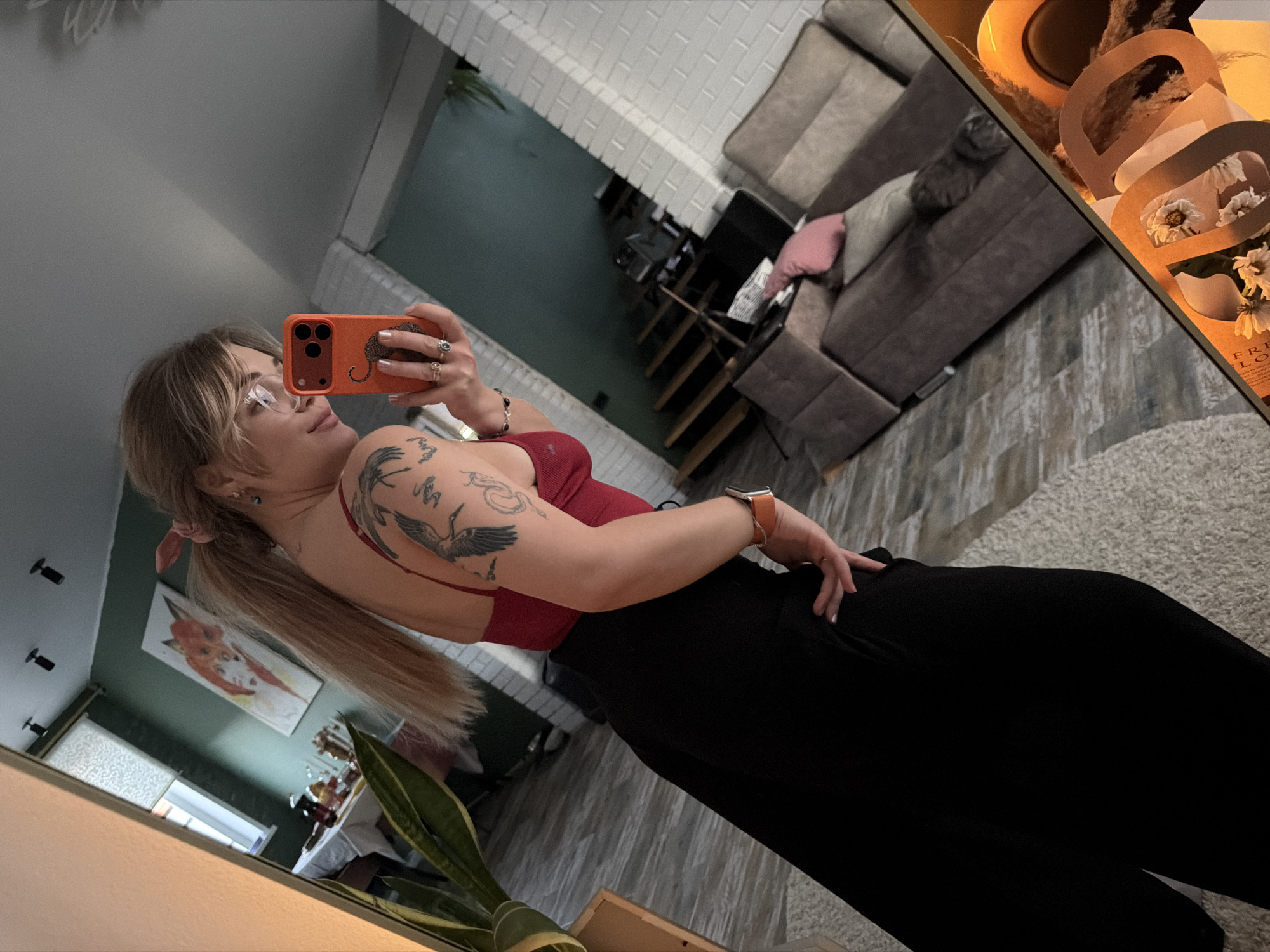 A woman is taking a mirror selfie indoors. She is wearing glasses, a red top, and black pants. She has blonde hair tied back with a pink scrunchie and tattoos on her arm. She is holding an orange smartphone with a graphic design. The background shows a living room with a gray sofa, stairs, and a painting of a fox on the wall. The image also shows part of a bouquet of flowers reflected in the mirror. The location appears to be in a residential setting. There's no clear indication that the photo was taken in the city of Большая Шильна, Russia.