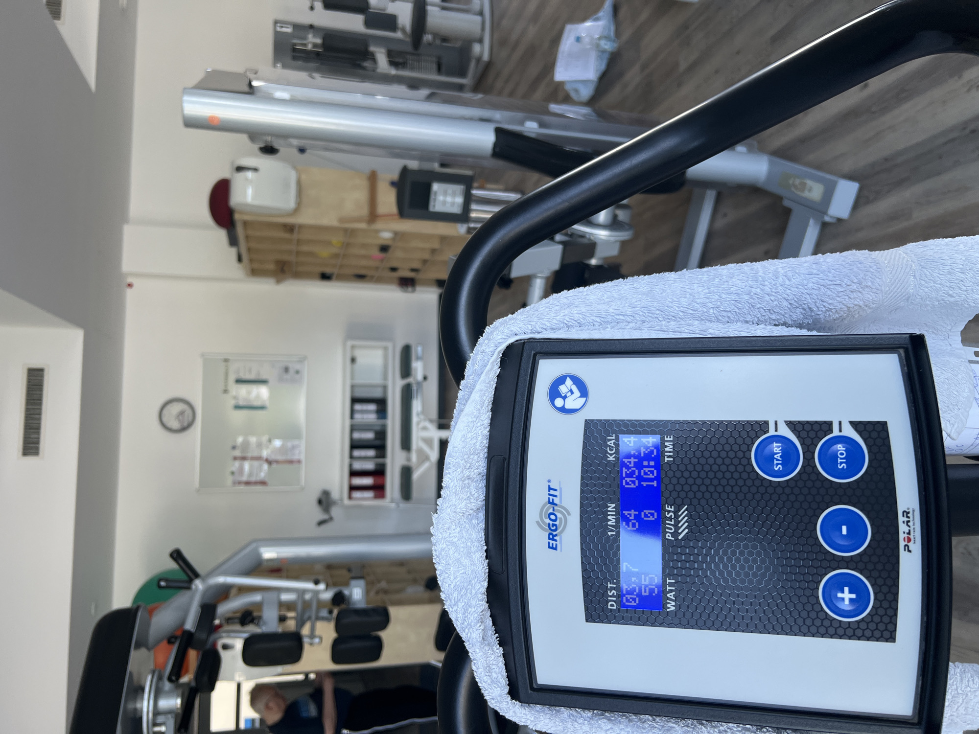 The image shows a close-up of a fitness machine console with a white towel draped over it. The console displays workout statistics, including distance, speed, calories burned, and heart rate. The brand "Ergo-Fit" is visible on the console, along with "Polar heart rate technology". In the background, the interior of a gym or rehabilitation center is visible, with other exercise equipment and storage shelves. A person in workout attire is partially visible in the left background, appearing to be using another machine. The environment appears to be well-lit and clean, with wooden floors. The scene is devoid of any explicit clues identifying it as being located in Teltow, Germany.