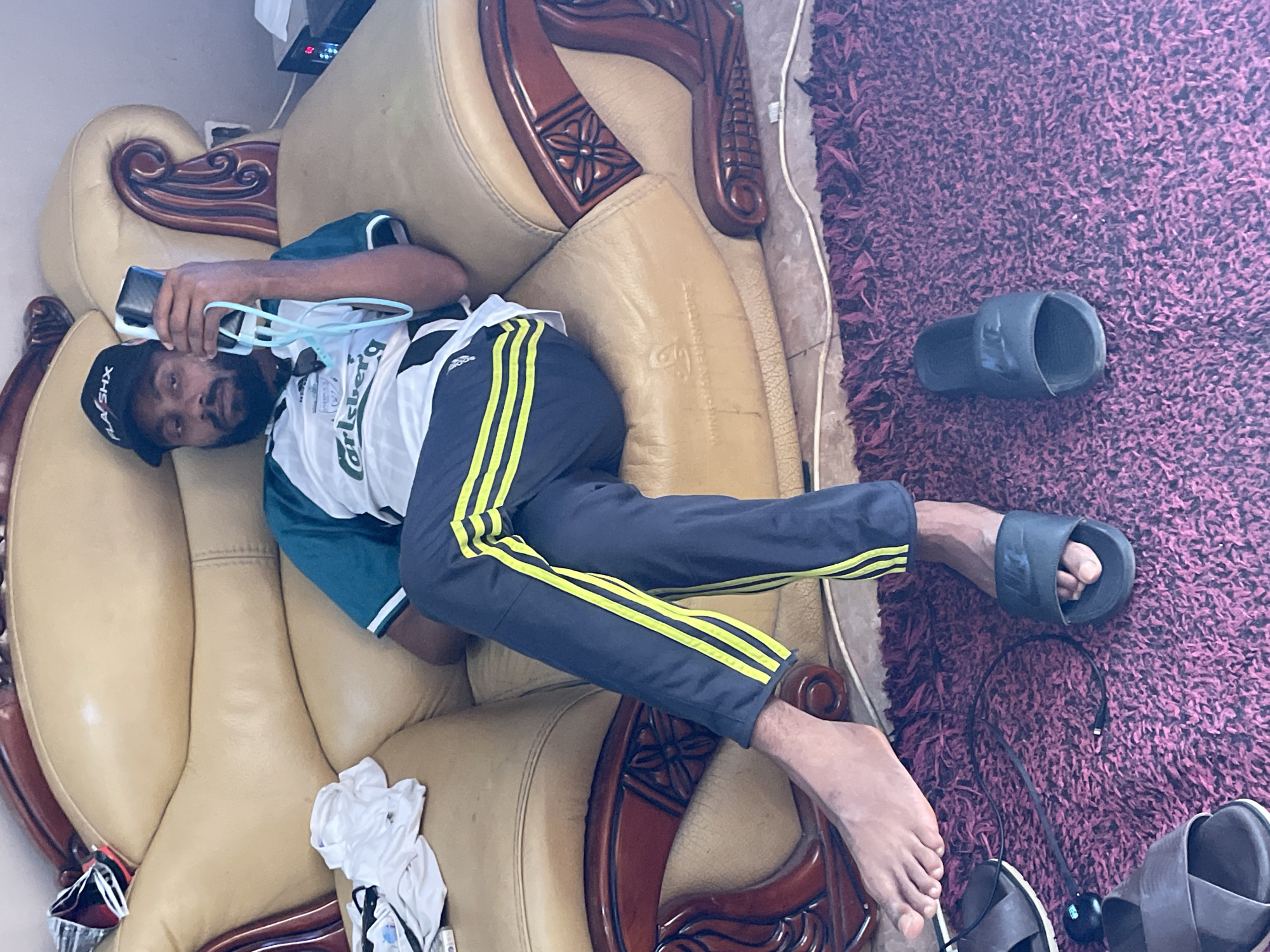 An indoor scene captures a man relaxing on a large, ornate sofa, likely in a living room in Jos, Nigeria. The dark-skinned man, appearing to be in his 20s or 30s, is lying on his left side, propped up on his arm. He has a short beard and mustache and is wearing a black cap with "FLASHX" or "FLASHE" written on it. His attire consists of a green and white short-sleeved top that clearly displays the "Carlsberg" logo in green, and dark grey track pants with bright yellow Adidas stripes down the sides.
In his right hand, he holds a black smartphone, seemingly engaged with its screen. Light blue-framed eyeglasses or a glasses holder are also near his hand. The sofa itself is upholstered in a light tan or beige material, featuring rich, dark carved wooden arms and frame. Adjacent to the sofa, a deep purple, shaggy rug covers the floor.
Several objects are scattered on the rug: a black charging cable, parts of what appear to be headphones, and multiple black slides or slippers, one of which clearly shows the "NIKE" logo. One of the man's bare feet rests on a wooden sofa arm, while the other foot, wearing a black slide, is on the rug. A crumpled white piece of clothing is also visible on the sofa near his feet. In the background, a white wall and a dark electronic device with glowing red numbers can be seen. The overall impression is one of casual relaxation during the daytime. Faint text "WINGSOVER FURNITURE" is visible on the sofa arm/cushion.