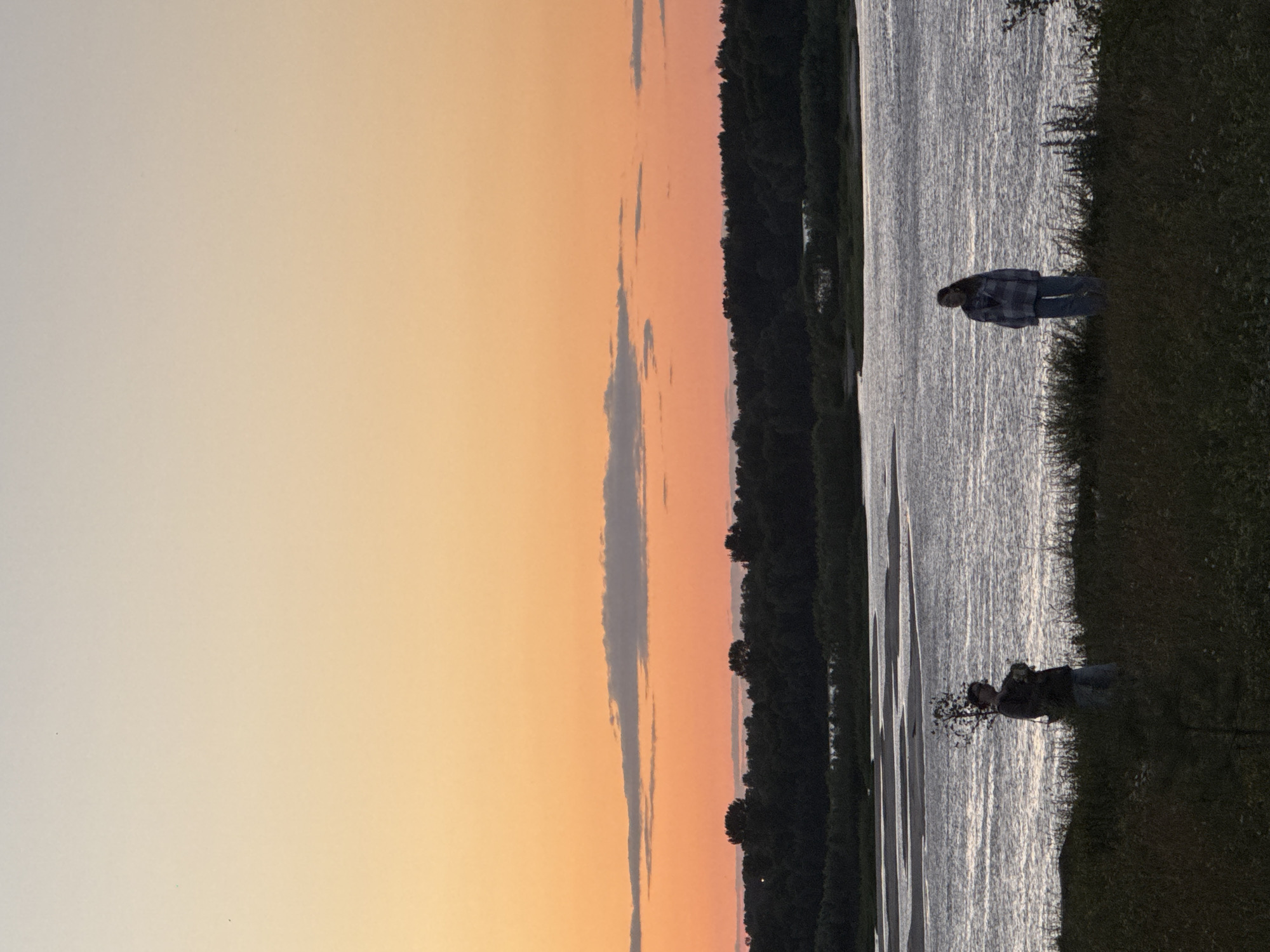 The media file captures a serene sunset scene on the banks of a body of water, likely a river or lake, in Nizhny Novgorod, Russia. The sky is a canvas of warm hues, transitioning from a soft, pale yellow at the top to a vibrant orange and pink as it meets the horizon. A few wispy clouds are scattered across the sky, adding depth to the atmospheric backdrop.
On the right side of the frame, a person is standing on the grassy bank, looking out towards the water. They are wearing a plaid shirt and jeans, and their silhouette is cast against the shimmering surface of the water. The water itself appears calm, reflecting the soft light of the setting sun, creating streaks of silver and white.
Further down the bank, near the water's edge, another figure is partially visible, appearing to be kneeling or bent over. This figure also seems to be enjoying the peaceful ambiance of the sunset. A line of dark trees forms a dense border along the far bank of the water, creating a natural contrast with the vibrant sky. The overall impression is one of tranquility and natural beauty, likely captured during the golden hour, a time often associated with a sense of calm and reflection. No visible text is present in the media file.