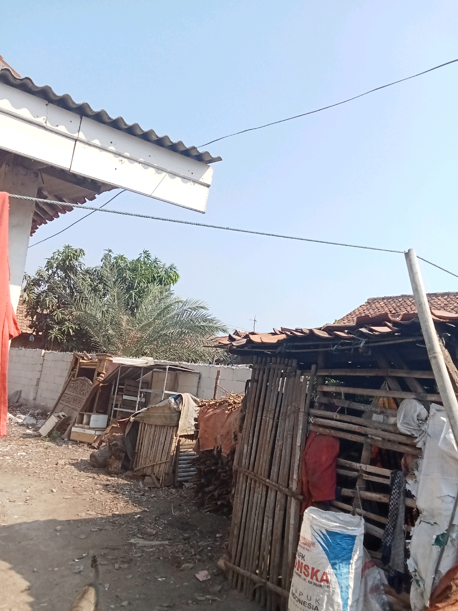 In Sumberreja, Indonesia, a rural outdoor scene unfolds under a clear blue sky. In the foreground, a dirt path is littered with debris, suggesting an unkempt or construction area. To the left, a white awning with a corrugated roof extends over a part of the scene, with a red fabric draped from its edge. Behind a low concrete wall, a date palm tree and other foliage are visible, hinting at a tropical climate. The central and right portions of the image are dominated by a makeshift structure constructed from bamboo poles and covered with corrugated metal roofing and fabric. This structure appears to be used for storage, as it contains various items including sacks of NPK fertilizer, rolled-up fabrics, and what looks like salvaged furniture or building materials. The overall impression is one of a modest, possibly developing, residential area with visible signs of everyday life and resourcefulness.
