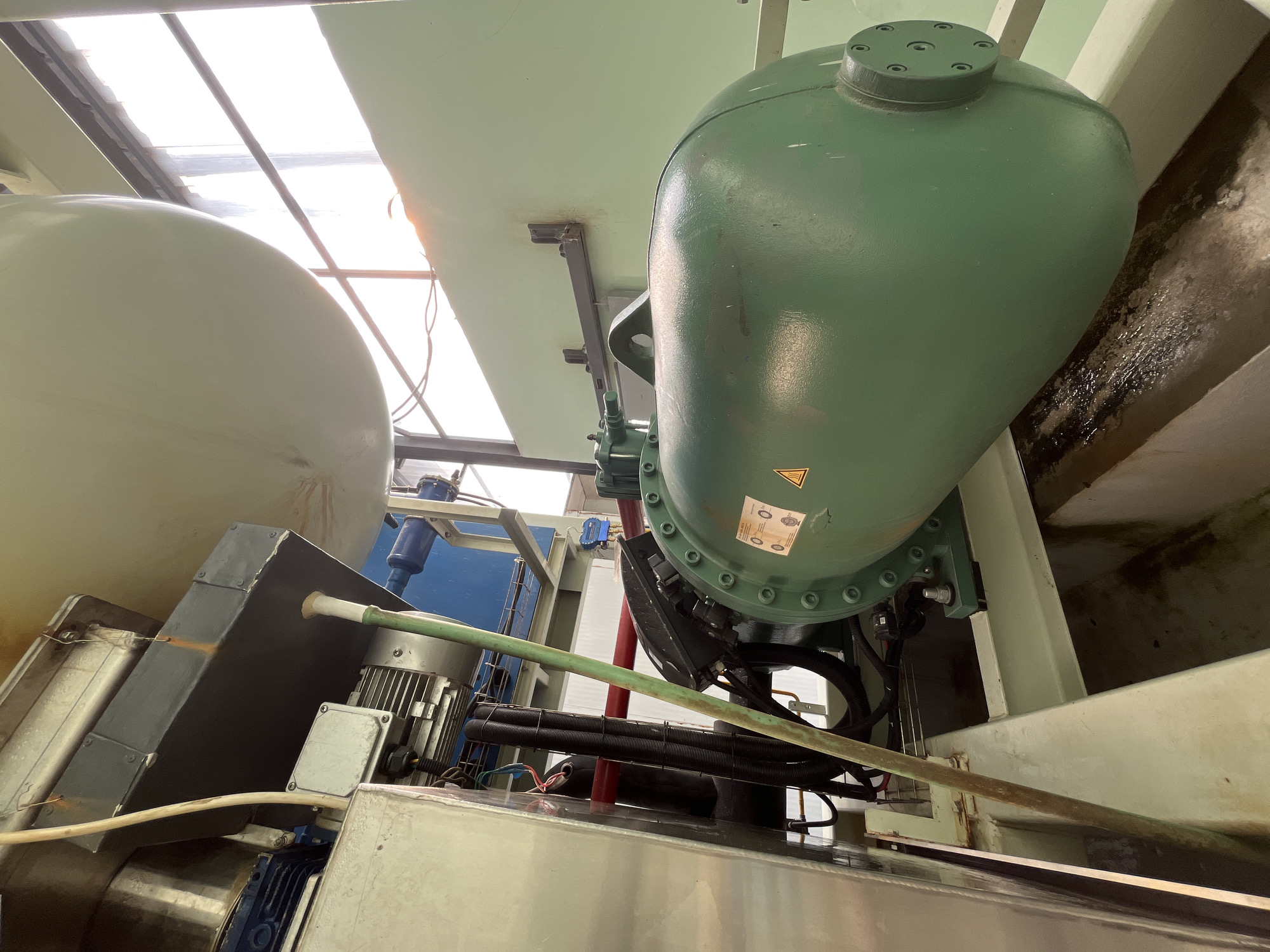 The image shows a close-up, low-angle shot of industrial machinery, likely part of a refrigeration or water treatment system, situated outdoors. The dominant feature is a large, cylindrical, green tank with bolted end caps, angled diagonally from the lower right to the upper left of the frame. Attached to the left side of this tank is a green, ribbed hose. Above and behind the green tank, a large, pale green, oval-shaped tank is visible. To the left of the green tank, a metallic structure with various pipes, electrical conduits, and a blue component are present. A blue motor is also visible in the upper left corner. The overall environment appears industrial, with visible metal framing and a glimpse of a window or skylight in the upper right, showing bright daylight, possibly suggesting a rooftop or elevated industrial facility. There are no people or animals visible in the image. The scene does not provide any specific indicators of its location in Pekalongan, Indonesia.