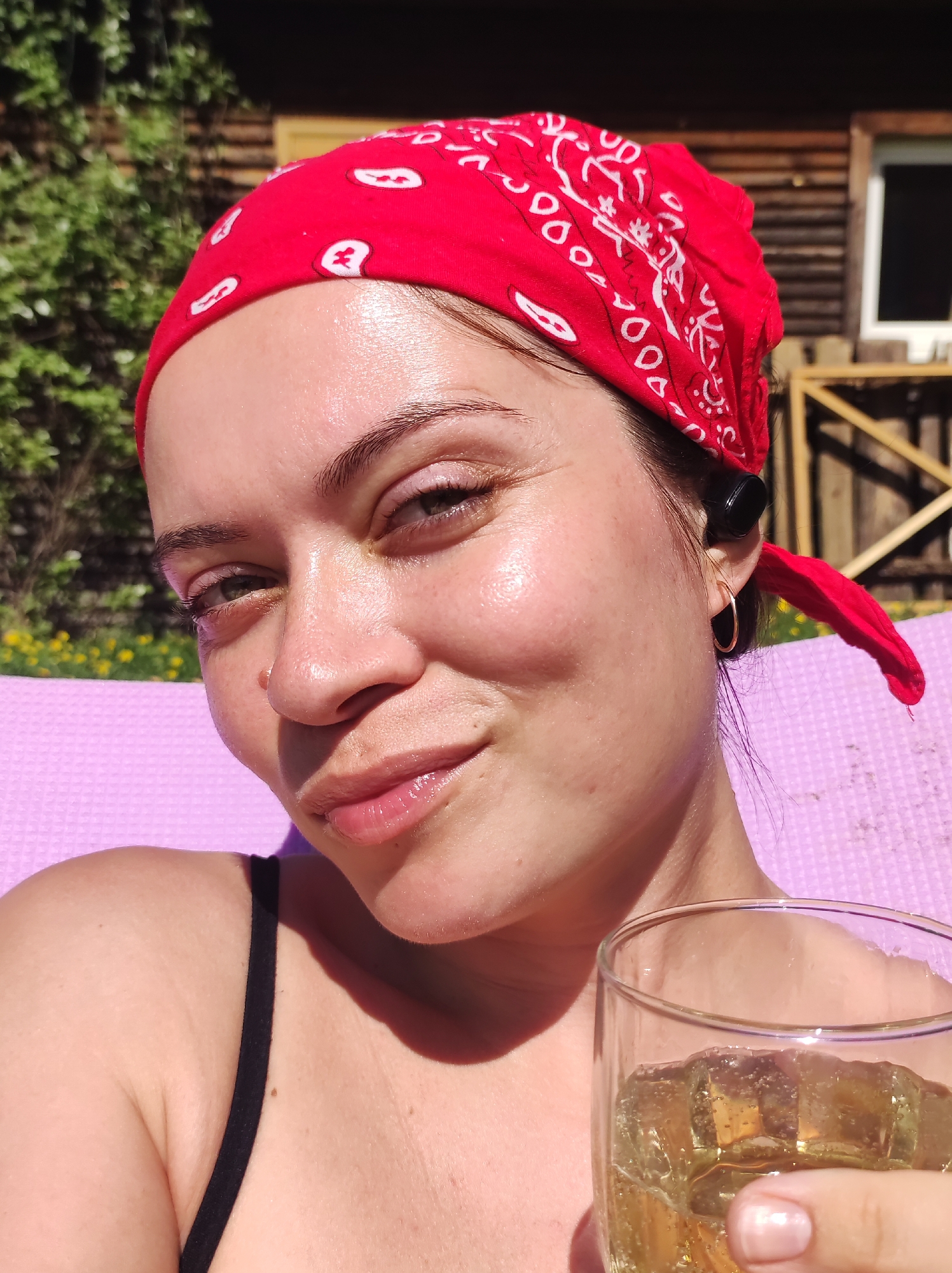 A close-up selfie shows a woman with fair skin, wearing a red bandana with a white paisley pattern. She has dark eyebrows and eyes that appear green or hazel. She is holding a glass of a pale yellow liquid with ice in her right hand. She is wearing small gold hoop earrings. In the background, there is a log cabin-style building and green foliage, indicating an outdoor setting. The person is sitting on a bright pink surface. The location in the city of Фоки, Russia cannot be determined from the visual data provided.