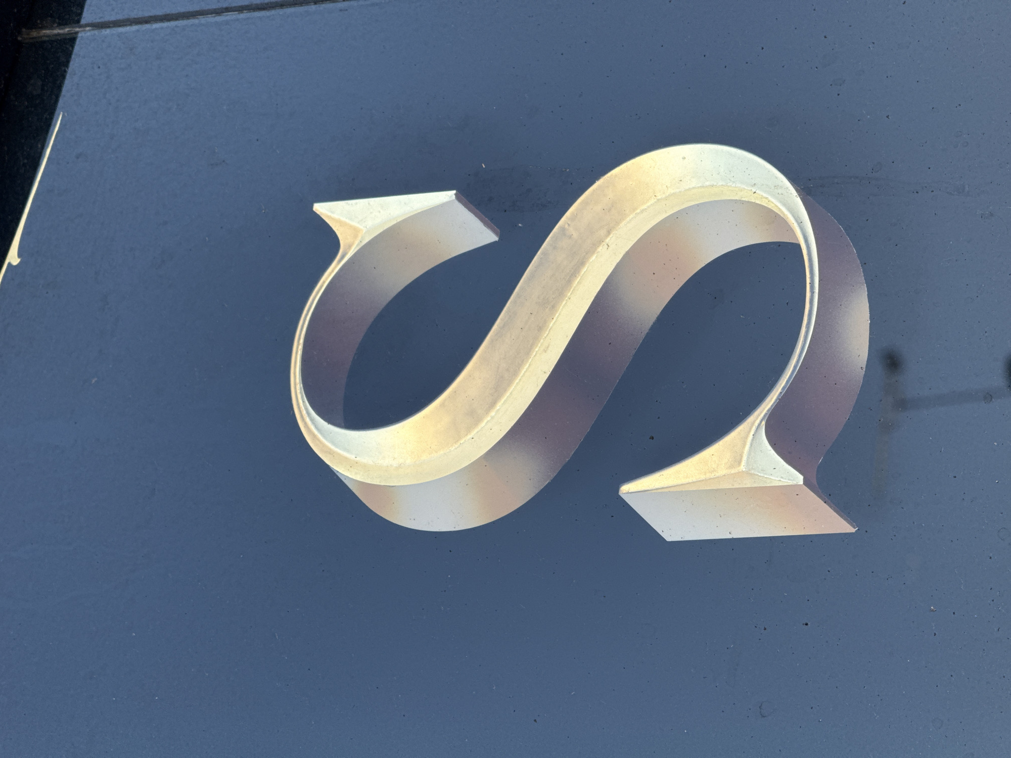 A close-up shot features a stylized, three-dimensional letter "S" mounted on a dark blue, slightly textured background. The "S" appears to be made of metal or a similar material with a metallic finish, exhibiting subtle variations in color, ranging from light gold to pale silver, likely due to lighting and reflections. The design incorporates flattened, angular ends. The background is a solid color, with minor imperfections and subtle surface texture visible. The composition is focused on the "S" with minimal surroundings visible. The location is Amersham, United Kingdom.