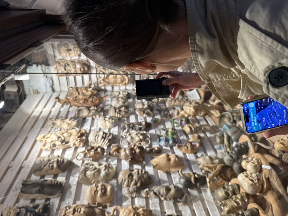 The image depicts an individual standing outside a shop window on Calle dei Stagneri o de la Fava in Venice, Italy. The person, viewed from behind and wearing a light-colored jacket, holds a small black digital camera in their right hand, aimed towards the display. Simultaneously, their left hand holds a smartphone displaying a map application that identifies the current location as "Calle dei Stagneri o de la Fava." The shop window behind the person contains a dense arrangement of numerous carved wooden artifacts, predominantly masks and decorative heads. These carvings vary in style, including detailed humanistic faces, mythical figures, and skulls, displayed on light-colored slatted wooden walls and shelves.