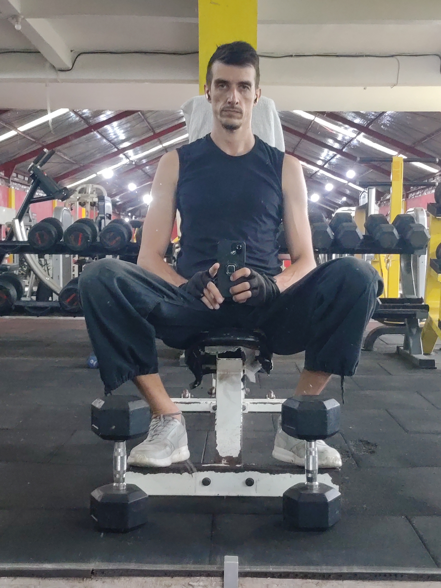 The image shows a man seated on a weight bench in a gym, located in Kedewatan, Indonesia. The man is wearing a black sleeveless shirt, dark pants, grey sneakers, and fingerless gloves. He is holding a black smartphone in his hands. Two dumbbells are positioned on the weight rack where his feet rest. In the background, various weight machines and dumbbells are visible. The gym appears to have a metal roof structure.