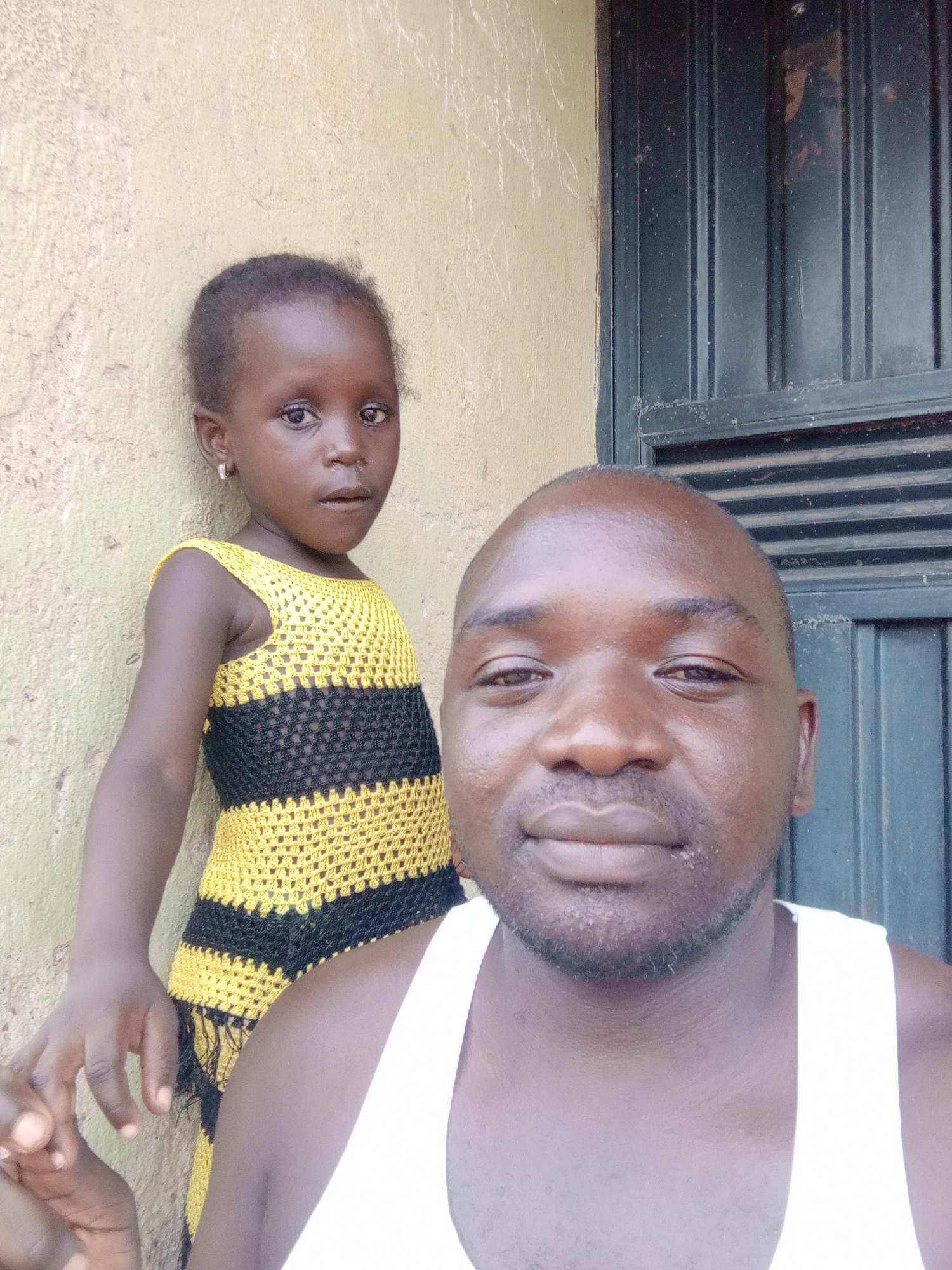 A medium close-up outdoor portrait features a man in the foreground and a young girl behind him, both with dark skin tones. The man, positioned on the right and front, has a shaved head, a short beard, and is wearing a white tank top. He looks directly at the camera with a neutral to slightly smiling expression.
Behind and to the left of the man, the young girl is visible, also looking towards the camera with a serious or curious expression. She has short, dark hair and wears small hoop earrings. Her attire is a distinctive yellow and black horizontally striped crocheted dress.
The background consists of a light-colored, possibly painted or stucco, wall on the left side, which shows some signs of wear, discoloration, and texture. To the right, behind the man and partly obscured, is a dark, possibly metal, door or gate with vertical panels and horizontal bracing. The lighting suggests it is daytime. No text is visible in the image. The context indicates the location is Akwanga, Nigeria.