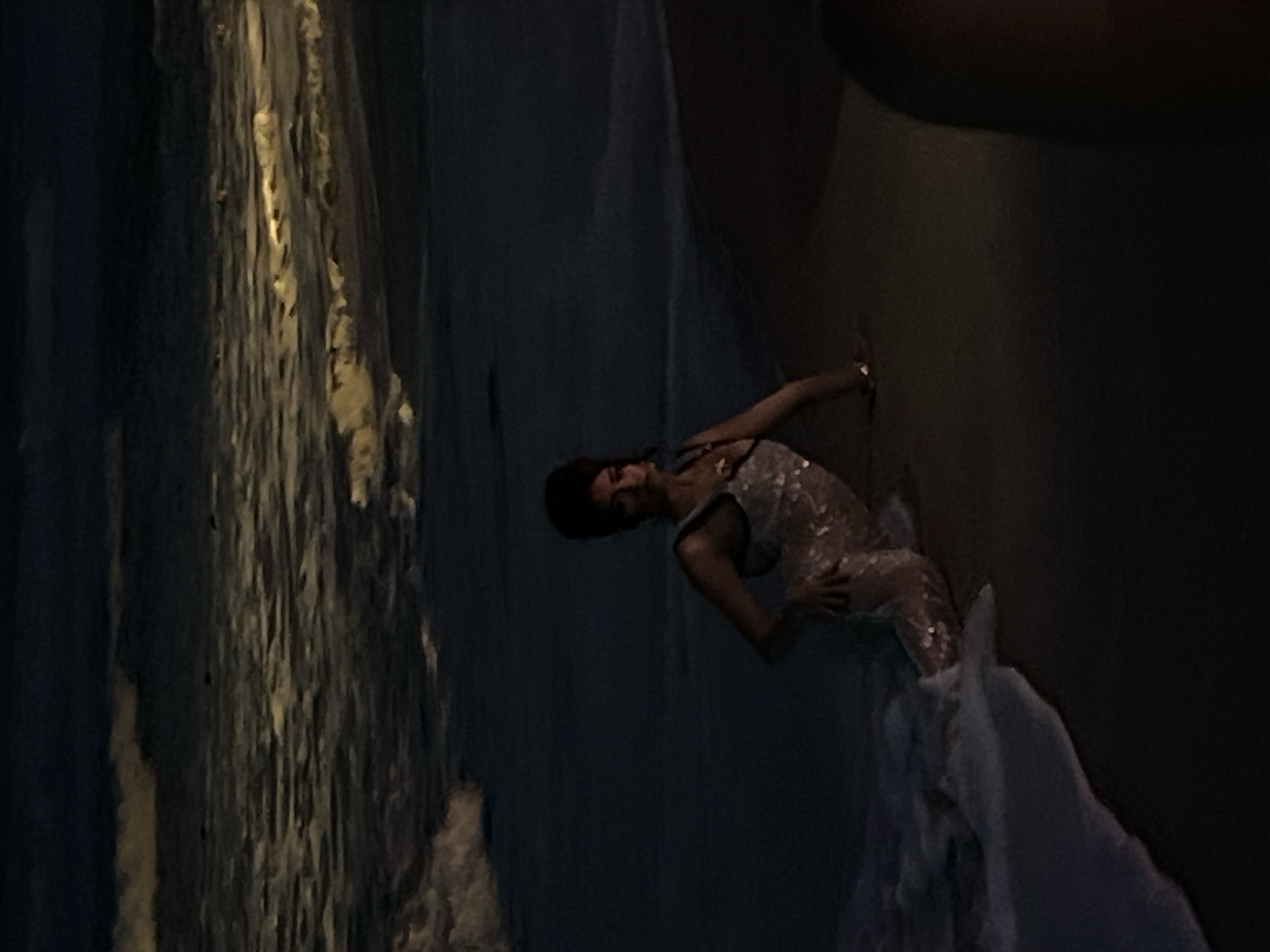 In an outdoor, low-light environment, a person is positioned on a sandy shore with waves gently lapping at their legs. The person, appearing to be female, is wearing a shimmering, form-fitting dress that extends to her knees. Her hair is styled in a voluminous updo, and she is leaning on her left hand, with her right hand resting on her thigh. The sea behind her is dark, with streaks of light reflecting on the choppy surface, suggesting artificial lighting or moonlight. The overall scene is dark and moody, with the water and sand dominating the composition.
