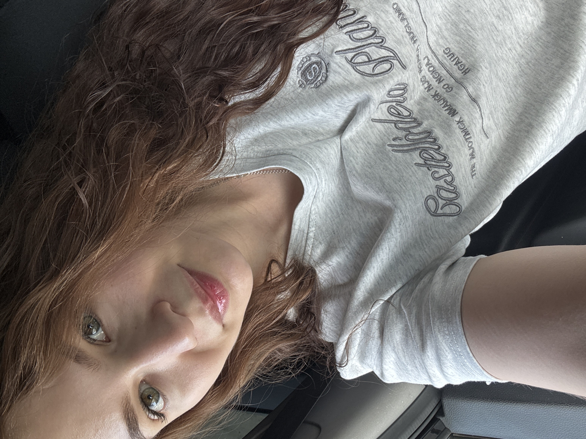 A young woman is photographed in a car. The photograph is shot from above, angled towards the woman's face. She has brown, wavy hair and green eyes. She is wearing a light gray, long-sleeved t-shirt with the words "Eastsider" and other text printed on it. Part of the car's interior is visible in the lower portion of the image, including a gray armrest. There is no specific location in the city of Chaikovsky, Russia depicted or implied by the image.
