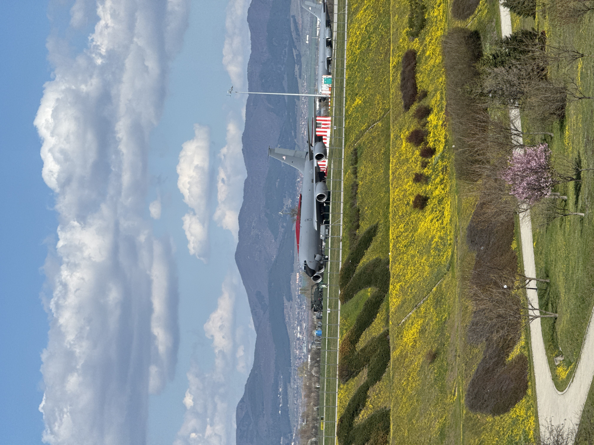 The image shows a passenger aircraft, likely parked, on what appears to be the tarmac of an airport. In the background are mountains. Adjacent to the tarmac is a field of bright yellow flowers. A walkway and flowering trees are also present.
Based on the type of aircraft, the mountains, and the presence of yellow fields, the image is likely depicting Sofia Airport in Bulgaria.