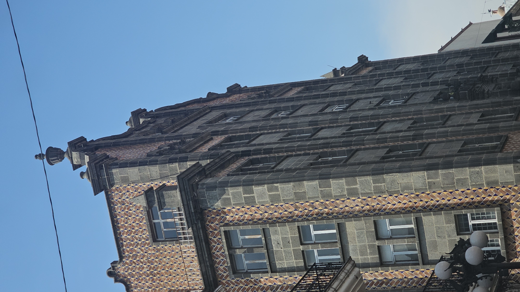 The image shows a portion of the facade of the Casa de los Muñecos in Puebla, Mexico. The building's architecture features a mix of gray stone and decorative Talavera tilework with geometric patterns. Several windows of various sizes are visible, some with ladders and security bars. The sky is clear blue. A power line is visible in the upper left corner.