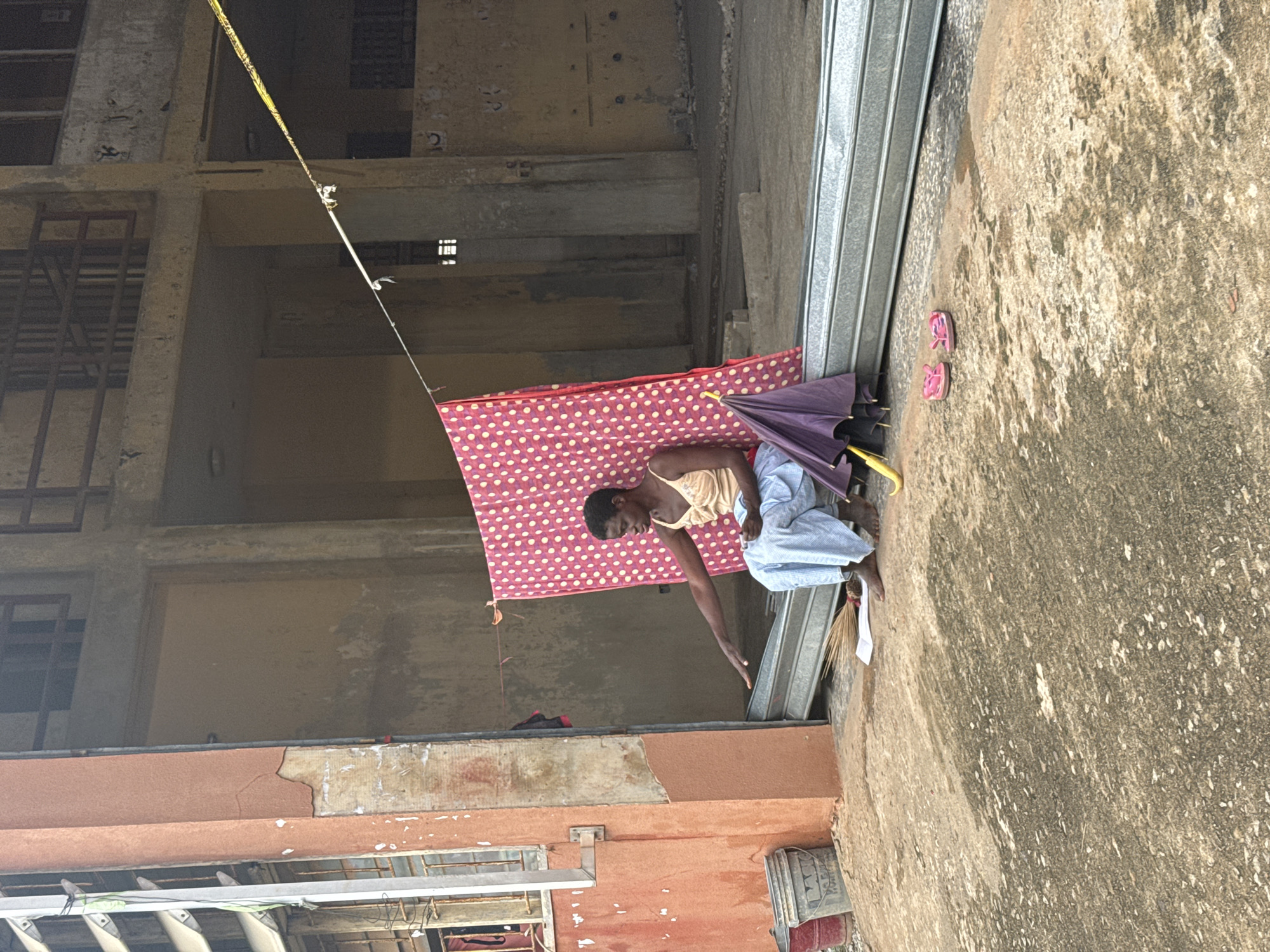 An outdoor scene captures a person crouching near a building, reaching down towards the ground. The image is oriented with the person on the right, bent over with their arm extended downwards. They are wearing a light-colored top and what appears to be a patterned blue and white skirt or wrap. Behind them, a red fabric with white polka dots is draped, possibly a mat or mattress, airing out. Next to the person, on the concrete ground, are a purple umbrella (partially open) and a pair of pink flip-flops. A small broom or brush is also visible near their feet.
The environment consists of weathered concrete buildings that appear somewhat dilapidated or under construction, with exposed beams and window frames. A yellow striped tape or rope stretches across the upper part of the frame, possibly for demarcation or support. The ground is a rough concrete surface. The lighting suggests it is daytime, possibly overcast or in a shaded area. The overall impression is that of a functional, lived-in space in a developing urban or semi-urban setting, consistent with the provided context of Ihiagwa, Nigeria. There is no discernible text visible in the image.