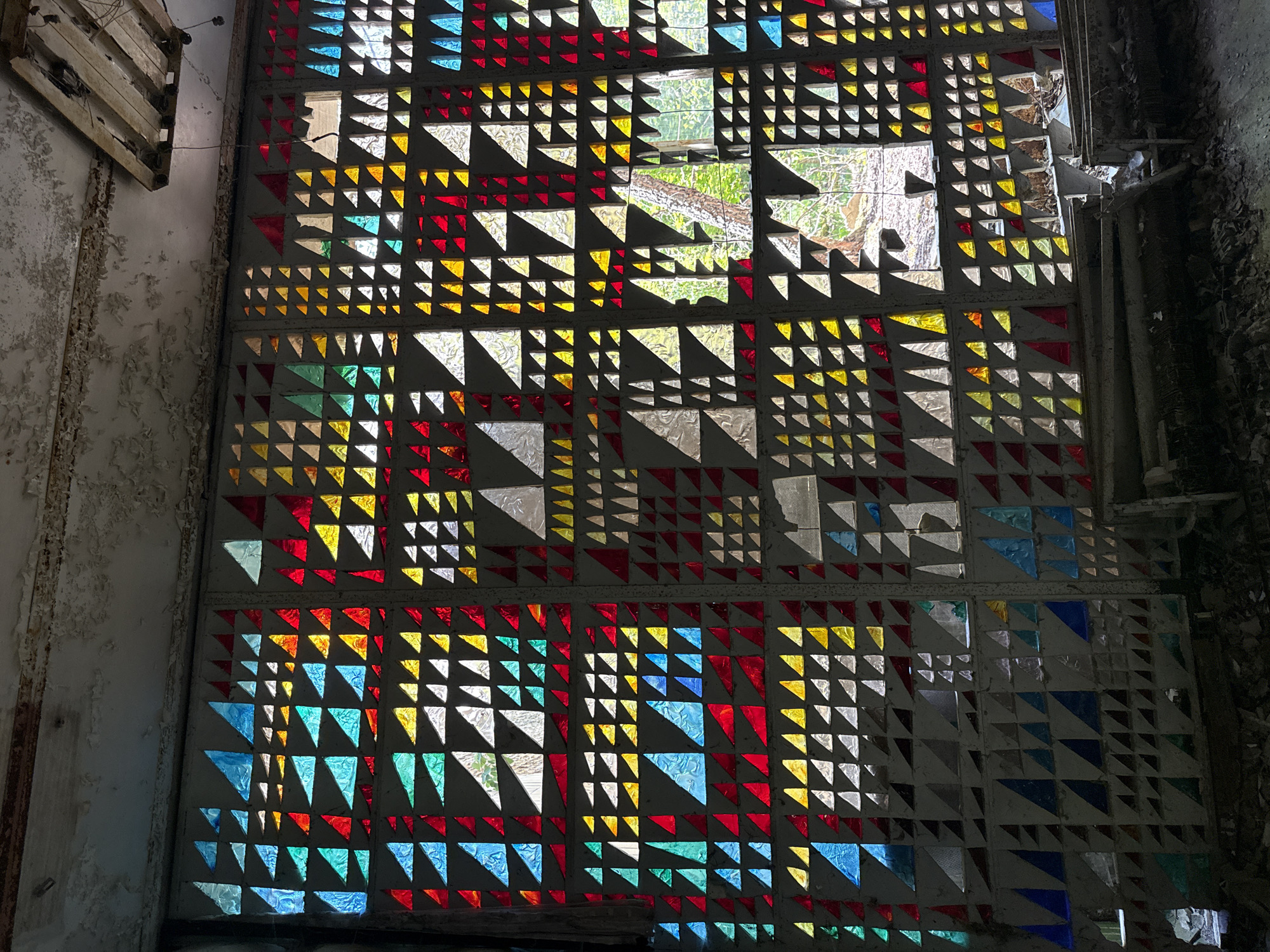 The image shows a large, geometrically patterned stained-glass window in a decaying building. The window is composed of squares, each containing smaller triangles of various colors: red, yellow, blue, green, and clear. Some squares are entirely open, revealing a glimpse of trees outside. The frame around the window is worn and damaged. The wall surrounding the window has peeling paint and visible deterioration. A broken, wooden structure is visible at the top left of the image. Given the unique style of the stained glass, it is highly probable that this image depicts the stained glass window located inside the "Avangard" Hotel in the city of Prypiat, Ukraine.