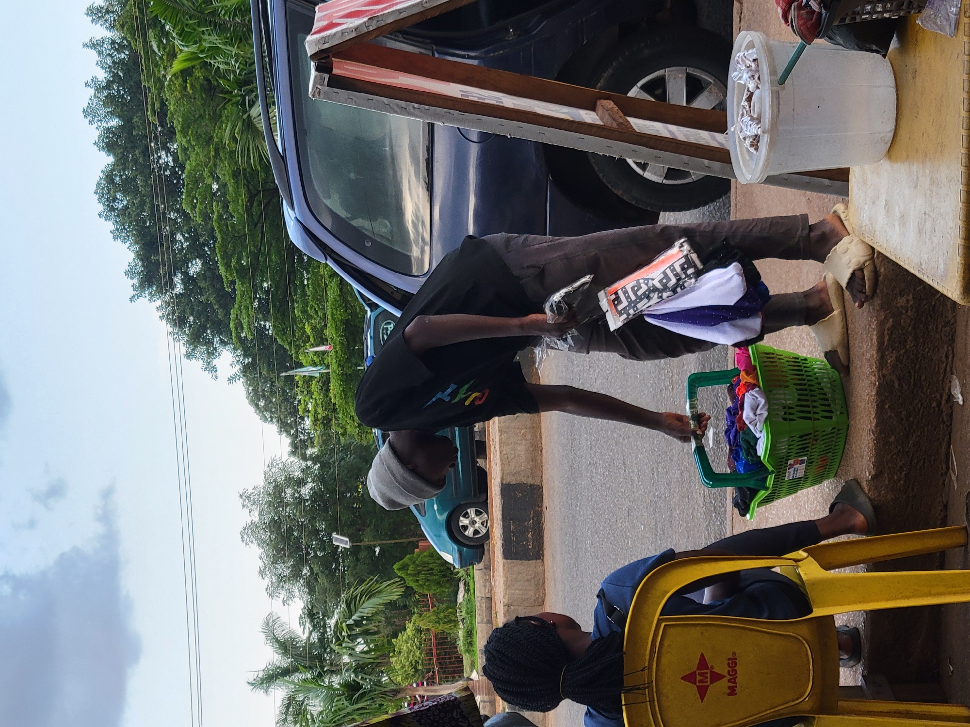 In Jos, Nigeria, a street vendor, wearing a grey beanie and a black t-shirt with colorful lettering, is interacting with a seated customer. The vendor is holding a plastic bag with folded clothes and a basket filled with various colorful fabrics. The customer, with braided hair, is sitting on a yellow chair bearing the "MAGGI" logo. Behind them, a dark blue van is parked, and a teal sedan is visible further down the road. The scene is set outdoors, with trees and a cloudy sky forming the background. A makeshift wooden stand with a white and red striped surface is also present.