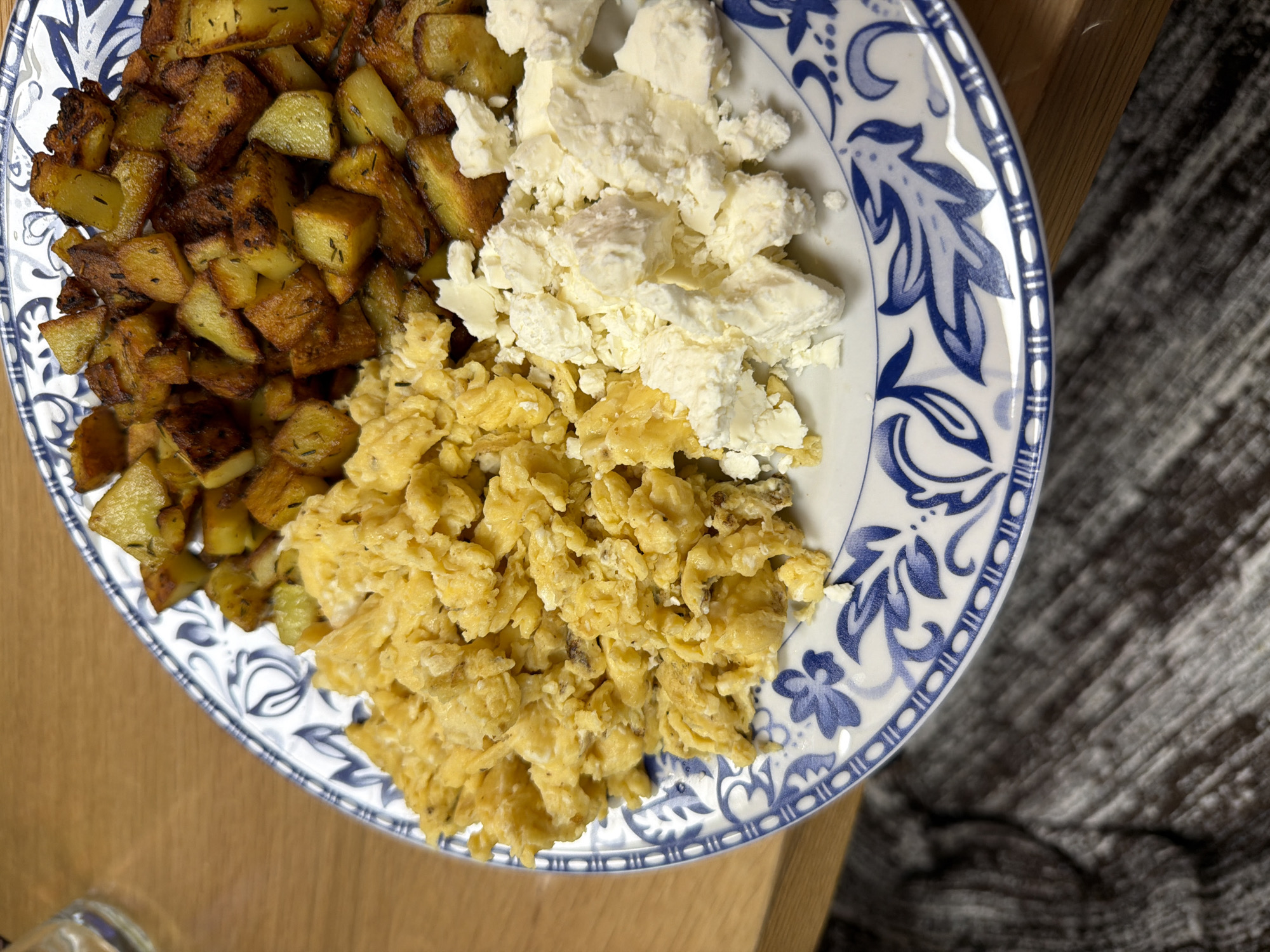 A plate with food on it is shown on a wooden table. The plate is round and has a blue and white floral design around the rim. The food consists of fried potatoes, scrambled eggs, and crumbled white cheese. The fried potatoes are browned and cubed. The scrambled eggs are yellow and appear slightly browned in places. The crumbled white cheese is in irregular chunks. The table is made of light-colored wood. A portion of a textured, gray surface is visible to the right of the plate. The location is not discernible based on the image.