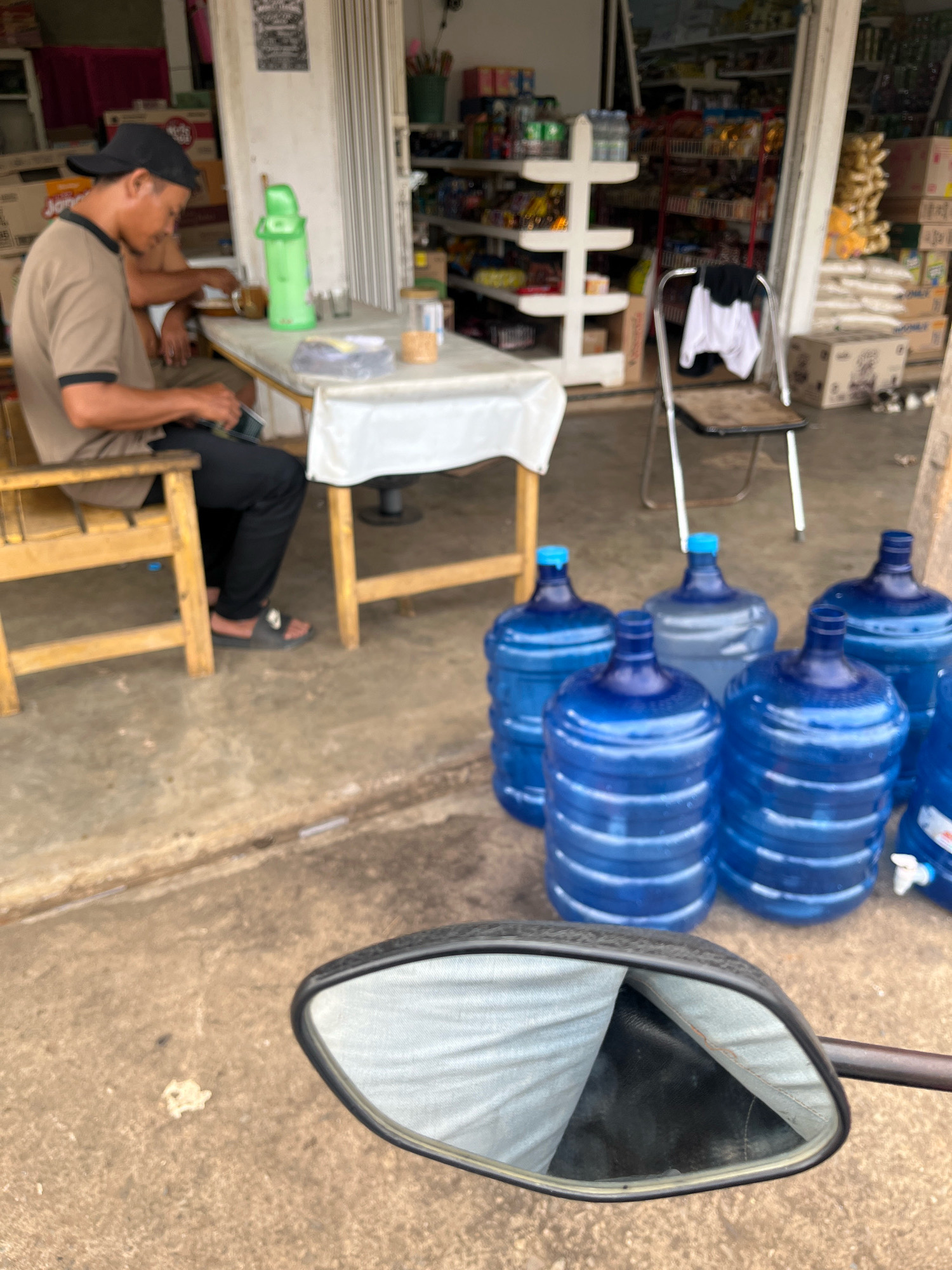 The image depicts a scene outside a small shop or warung in Muara Dua, Indonesia. In the foreground, a motorcycle's side mirror is visible, reflecting a person wearing a light blue shirt and black pants. Behind the mirror, a person wearing a tan t-shirt and a black cap is seated at a table, looking down at their phone. On the table are a green thermos, a glass, and a small container. The table is covered with a white cloth. To the right of the table, there are several large, blue water cooler jugs lined up on the ground. Behind the jugs and the table, the interior of the shop is visible, with shelves stocked with various items. A folding chair with a black and white garment draped over it stands near the shop entrance. The ground is a concrete surface. The overall impression is that of a typical roadside establishment in a warm climate.