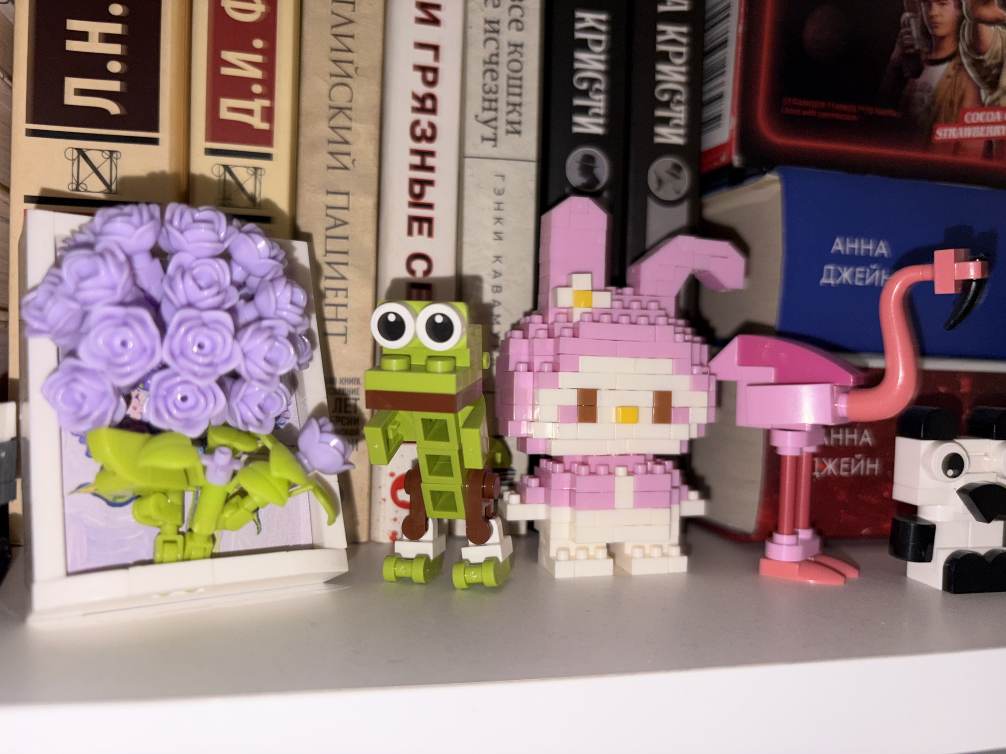 The image captures a vibrant display of various block-built figures and books on a shelf in Perm, Russia. The scene features a whimsical collection of miniature characters standing in front of several upright books, predominantly in Cyrillic script.
From left to right, the display includes a framed arrangement of purple plastic flowers, possibly hydrangeas or roses, depicted as if in a window. Next to it stands a green frog-like figure with prominent white eyes and black pupils. Following the frog is a pink and white character, identifiable as My Melody from Sanrio, constructed from small building blocks. A pink flamingo figure with dark pink legs and beak extends towards the right. Partially visible on the far right is a black and white figure, likely a panda, also made of building blocks.
Behind these figures, several books are arranged. Visible text on their spines includes "Л.Н.", "Д.И. Ф", "ГЛИЙСКИЙ ПАЦИЕНТ" (likely "The English Patient"), "И ГРЯЗНЫЕ СЕ", "все кошки исчезнут" (all cats will disappear), "ГЭНКИ KABAM" (Genki Kawamura), and multiple instances of "КРИСТИ" (likely Agatha Christie). There's also a red box featuring "STRANGER THINGS" and "COCOA STRAWBERRY", and a blue book with "АННА ДЖЕЙН" (Anna Jayne) visible on its spine. The setting is an indoor shelf, suggesting a personal collection or decorative display. No specific activity is occurring, and the time of day is not discernible.