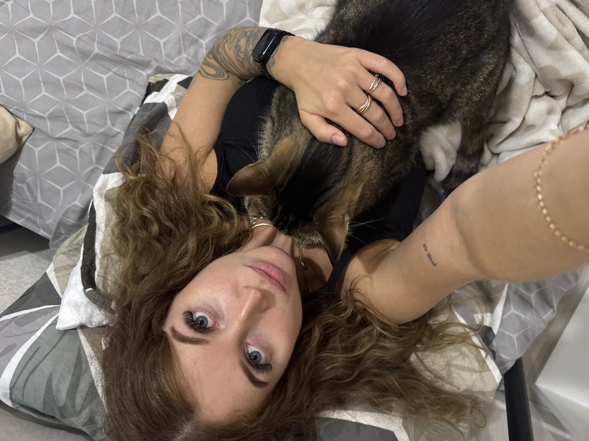 An overhead shot captures a woman lying on a bed with a tabby cat on her chest in what appears to be Chaikovsky, Russia. The woman has long, curly brown hair fanned out around her head and bright blue eyes gazing directly at the camera. She wears a black top. Her right arm, adorned with a large, intricate tattoo sleeve and a black smartwatch, is bent with her hand gently resting on the cat's back, multiple gold rings visible on her fingers. The cat, featuring dark brown and black striped fur, rests its head near her neck, appearing to nuzzle her. A delicate gold necklace is visible around her neck. Her left arm extends towards the bottom right of the frame, revealing a small cursive tattoo on her inner forearm and a thin gold chain bracelet on her wrist. The bedding consists of a gray and white geometric patterned duvet cover and a plain white blanket. The scene conveys a relaxed and affectionate moment between the woman and her pet indoors, likely during the daytime or evening.