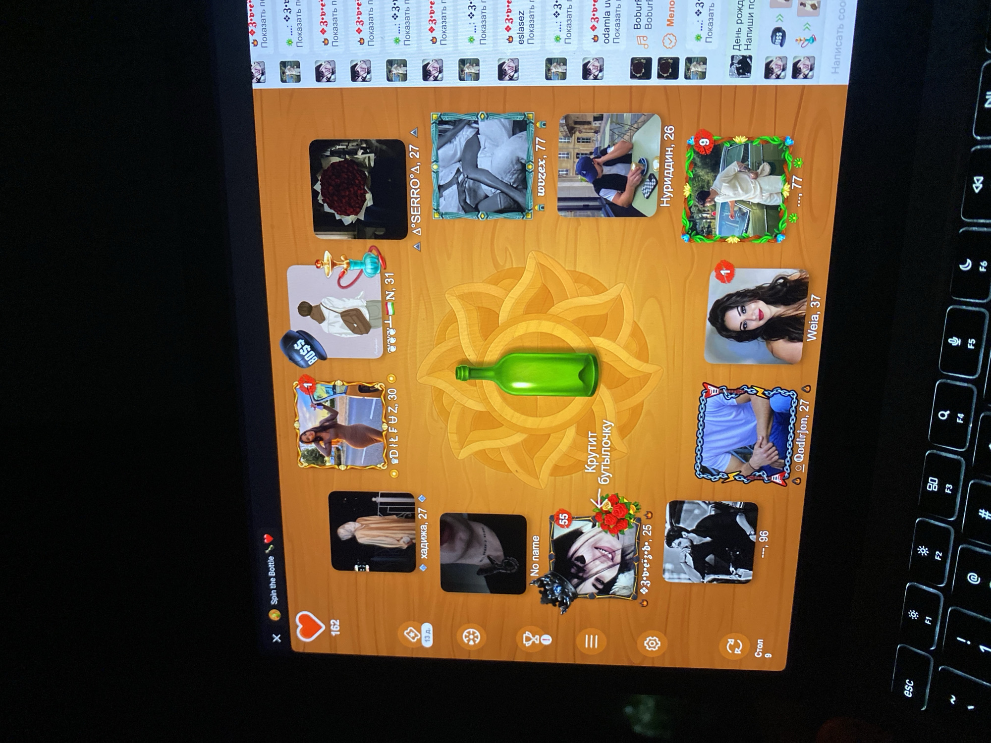 The image depicts a screenshot of a game interface, likely a "spin the bottle" application, displayed on a laptop screen. The game is set against a wooden background with various player avatars and game elements. In the center, a large green bottle graphic is prominent, suggesting the game's core mechanic.
To the left of the bottle, there are navigation icons and a heart icon with the number 162. Below this, a list of icons includes a trophy and a hamburger menu. Further down, a "Стол 9" (Table 9) is indicated, along with a refresh icon.
Player avatars are arranged around the central bottle. These avatars include profile pictures of individuals with their associated usernames and ages. Examples include "хаджика, 27", "No name", "Weia, 37", and "Нуриддин, 26". Some avatars have numerical badges, such as "55" and "1".
On the right side of the screen, a scrolling list of notifications or messages is visible, displaying user names, timestamps, and icons.
The laptop's keyboard is visible at the bottom of the image, indicating the device on which the game is being played. The overall environment appears to be indoors, with the glow of the screen illuminating the keyboard.
The request to include the city of Khankurgan, Uzbekistan, is not supported by the visual information in the image. The image is a digital interface and does not contain any geographical markers or contextual clues that would place it in a specific city or country.