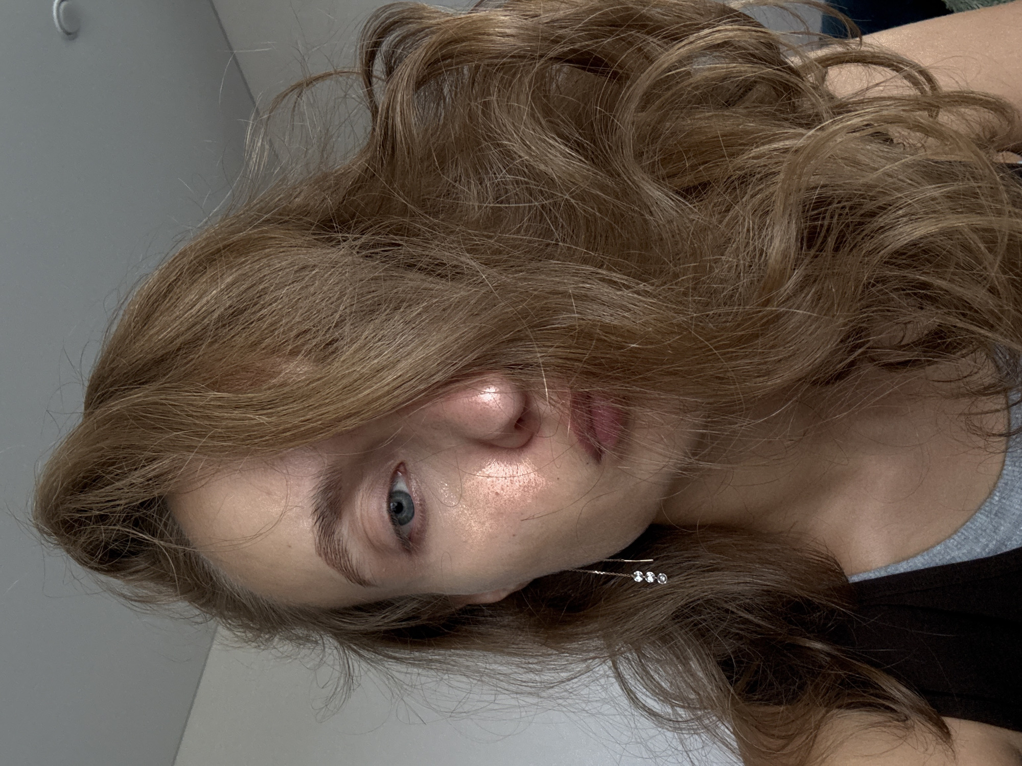 A close-up, waist-up portrait of a young woman is captured against a plain, light-colored wall. She has medium-length, wavy brown hair that is styled to obscure the right side of her face and fall across her left eye. Her left eye, which is visible, is blue. Her skin appears fair with a subtle sheen, and she is wearing a delicate silver earring with three small, clear stones dangling from her left ear. She is dressed in a dark top layered over a gray tank top. The setting is indoors, with the ceiling visible in the upper portion of the frame, and the lighting suggests it is daytime. There are no identifiable features of the city of Подшивалово, Russia in the image.