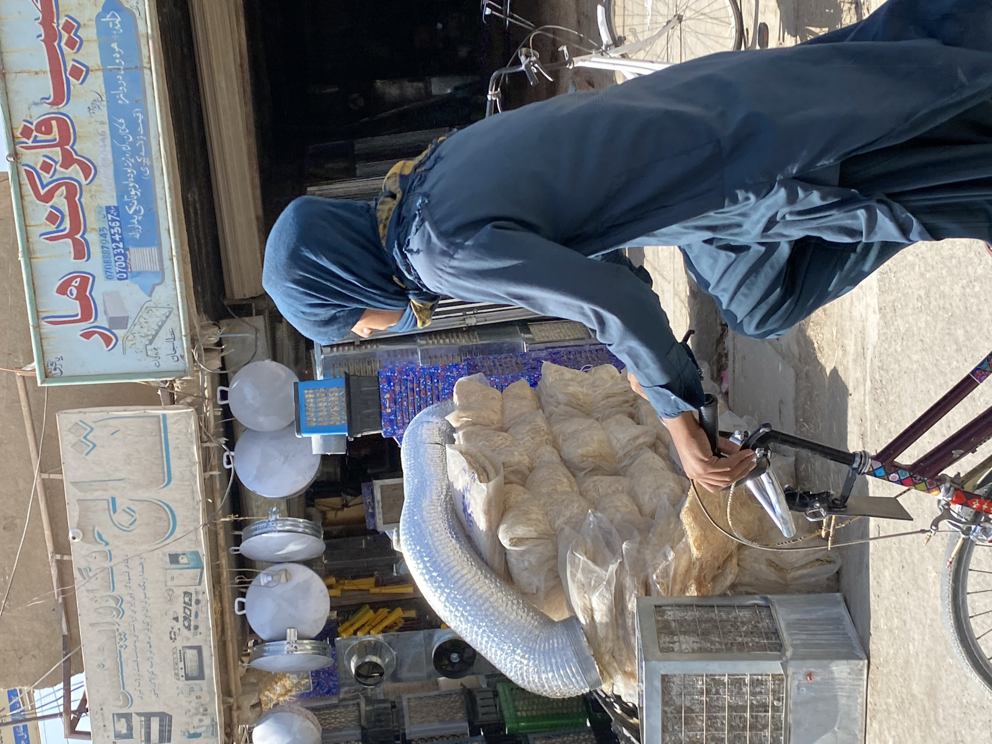 The image depicts a street scene in Kandahar, Afghanistan, with a young person on a bicycle. The individual is wearing a dark blue hijab and a matching long tunic and pants, common attire in the region. They are holding the handlebars of a bicycle, which appears to be older and has decorative elements on the frame.
The background reveals a marketplace setting. Prominent signage, written in Pashto, advertises products and services. One sign, with red and blue lettering, prominently displays "صبيب فلزکند هار" (Sabib Filzkind Har). Another sign to the left, featuring illustrations of appliances, reads "و دواريس" (wa Dawaress). Various goods are displayed, including what appear to be air coolers, large stacks of bundled materials wrapped in plastic, and metallic cylindrical ducts. The overall environment suggests a busy commercial area with an outdoor display of goods. The lighting indicates it is daytime, with shadows cast on the ground.