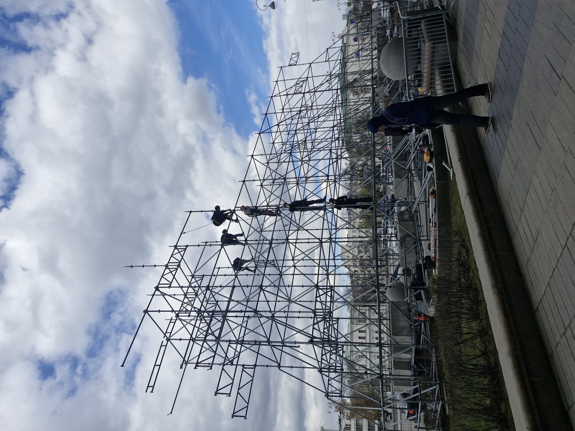 A large, intricate metal scaffolding structure dominates the right side of the image, extending upwards towards a partially cloudy sky. Several individuals, likely workers, are actively engaged on different levels of the scaffolding, appearing to climb and work on its assembly or modification. They are silhouetted against the bright sky, some wearing what appear to be work safety gear like helmets.
To the left of the scaffolding, a paved sidewalk or plaza is visible. A person in dark attire, possibly wearing a beanie, stands on this paved area, seemingly observing the scaffolding and the activity on it. Metal barriers or temporary fencing run along the edge of the paved area, suggesting a designated zone or ongoing event setup. Beyond the sidewalk, a strip of sparse, bare bushes lines a small grassy area. In the background, urban buildings with light-colored facades are discernible, hinting at a city environment. The sky above is a mix of bright white and grey clouds interspersed with patches of clear blue. The scene is illuminated by daylight. The image appears to be rotated 90 degrees counter-clockwise. There is no visible text. The setting is an urban environment, specifically in Yekaterinburg, Russia.