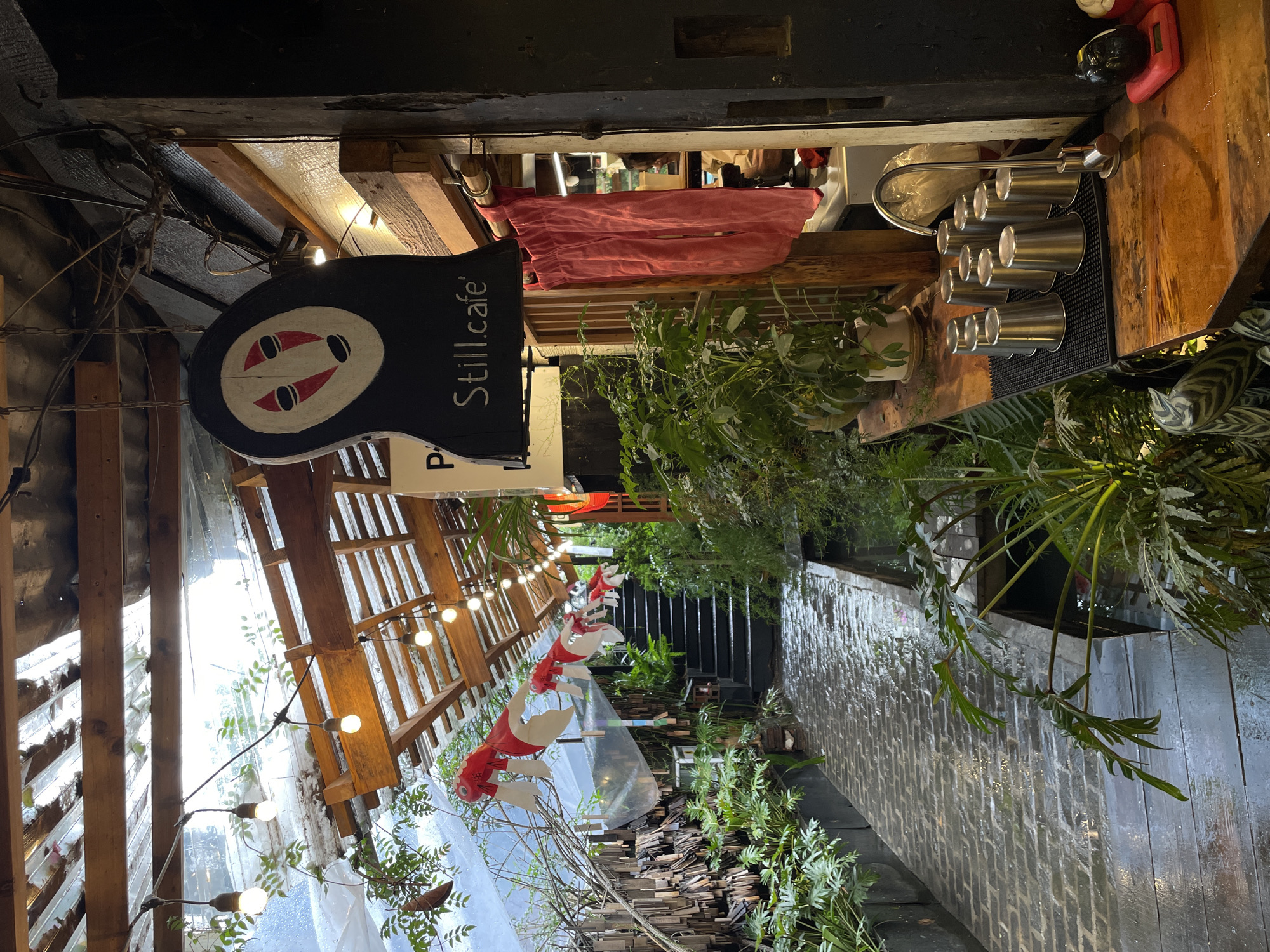 This is an exterior view of the Still Cafe in Lâm Viên - Đà Lạt, Vietnam. The cafe's entrance is visible with a hanging sign featuring a cartoon face and the cafe's name. The cafe is decorated with greenery, including plants and trees lining the walkway leading to the entrance. There are strings of lights hanging overhead and paper koi fish decorations. The walkway is paved with stone and there is a waterfall feature on one side. Inside the cafe, there are espresso preparation tools. Above, there's a red piece of fabric, possibly a garment.