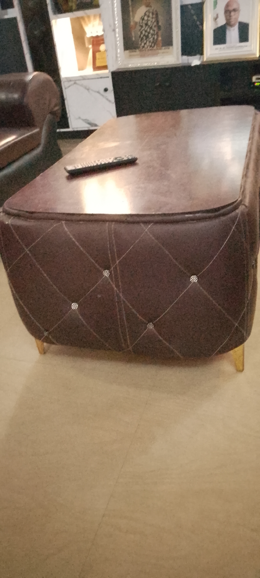 This is a medium-angle, eye-level shot of a coffee table in what appears to be a living room or office space. The coffee table is upholstered in dark brown material with a diamond-stitched pattern, accented with small, reflective embellishments at the intersection of the stitches. The table has a glossy, dark brown top and rests on four short, golden-colored legs. A black remote control is placed on the surface of the table. In the background, a tall, white cabinet with some framed pictures and objects can be seen. One framed picture shows a person in traditional attire, and another shows a person in formal wear wearing glasses. The overall impression is of an indoor domestic or professional setting, possibly in Gboko, Nigeria, judging by the prompt's context.