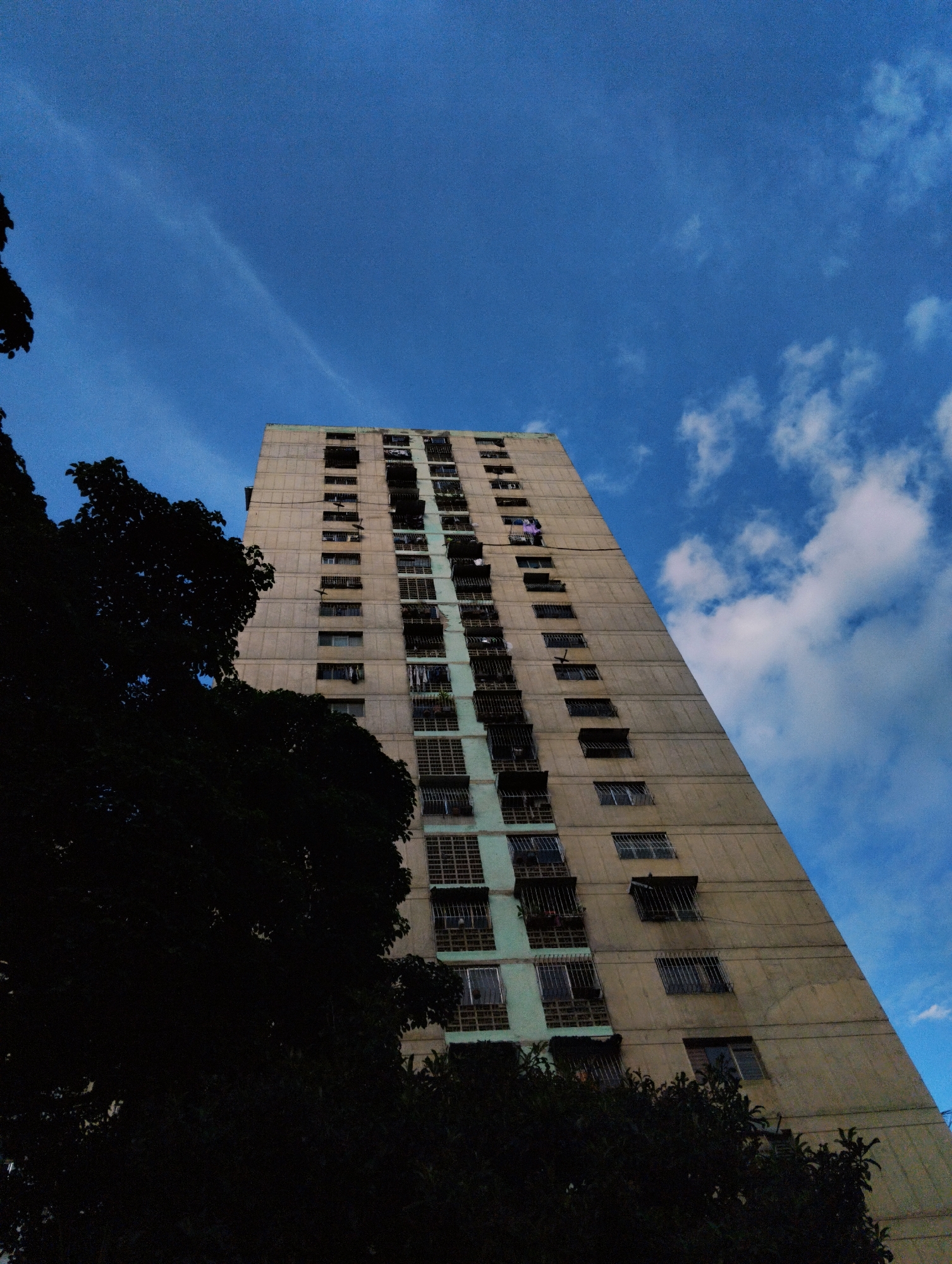 A tall, concrete apartment building, characteristic of Venezuelan Brutalist architecture, is captured from a low angle in Caracas. The building, with its repetitive grid of windows and balconies, dominates the frame, partially obscured by the dark silhouette of lush, leafy trees in the foreground. The sky above is a vibrant blue, scattered with thin, wispy clouds, suggesting it is either early morning or late afternoon. Laundry hangs from some of the balconies, a common sight in urban Venezuela, adding a touch of domestic life to the imposing structure. The overall impression is one of urban density against a natural sky.