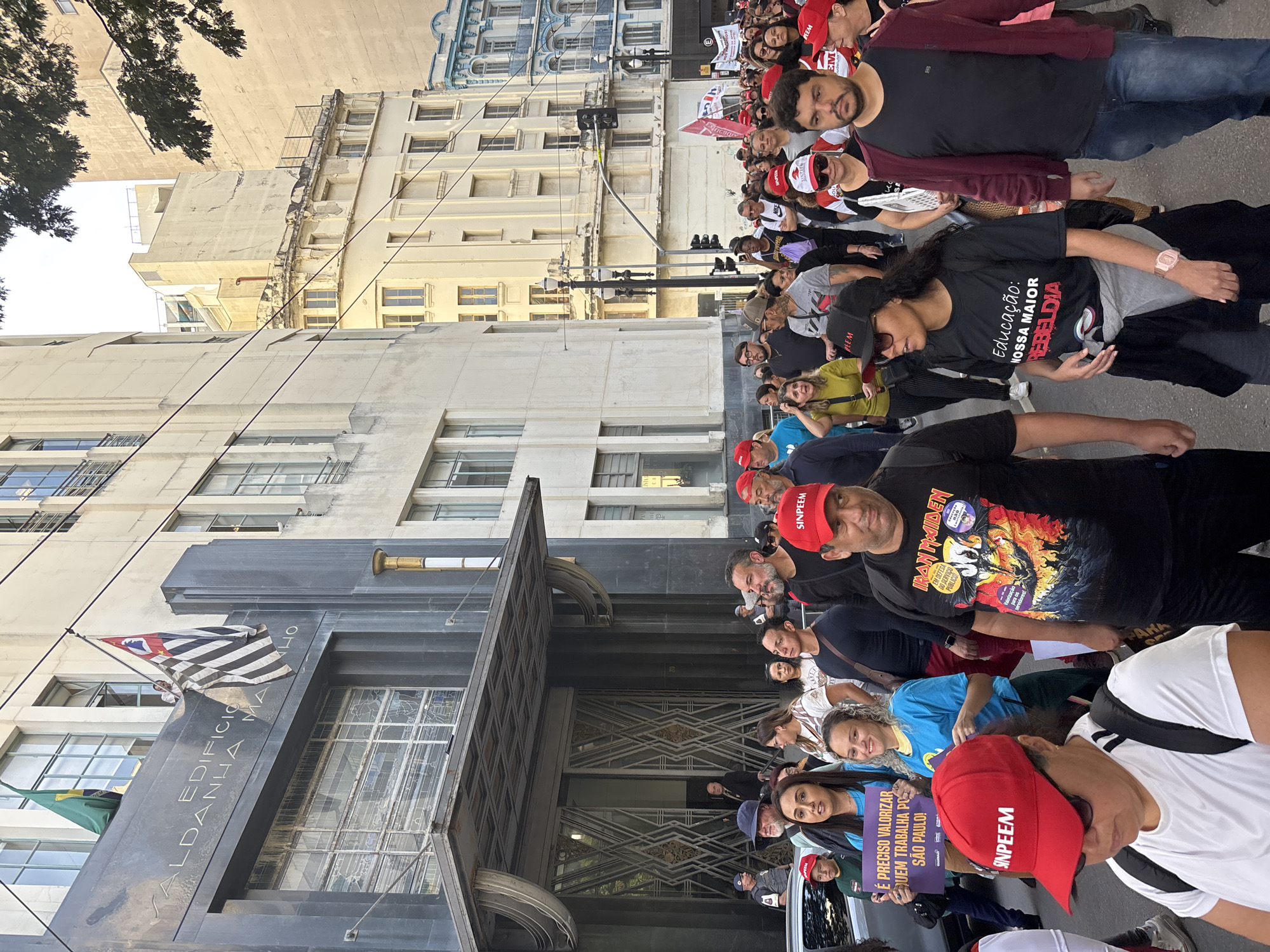 A crowd of people is standing in front of the Edifício Saldanha Marinho in São Paulo, Brazil. The flags of São Paulo and Brazil are hanging from the building. Many of the people in the crowd are wearing red hats with the word "SINPEEM" printed on them. Some are holding signs that say "É PRECISO VALORIZAR QUEM TRABALHA PO SÃO PAULO!" and "Educação: NOSSA MAIOR REBELDIA." One person is wearing a shirt with an Iron Maiden graphic on it. The street and buildings around them appear to be busy, suggesting a public demonstration or protest.