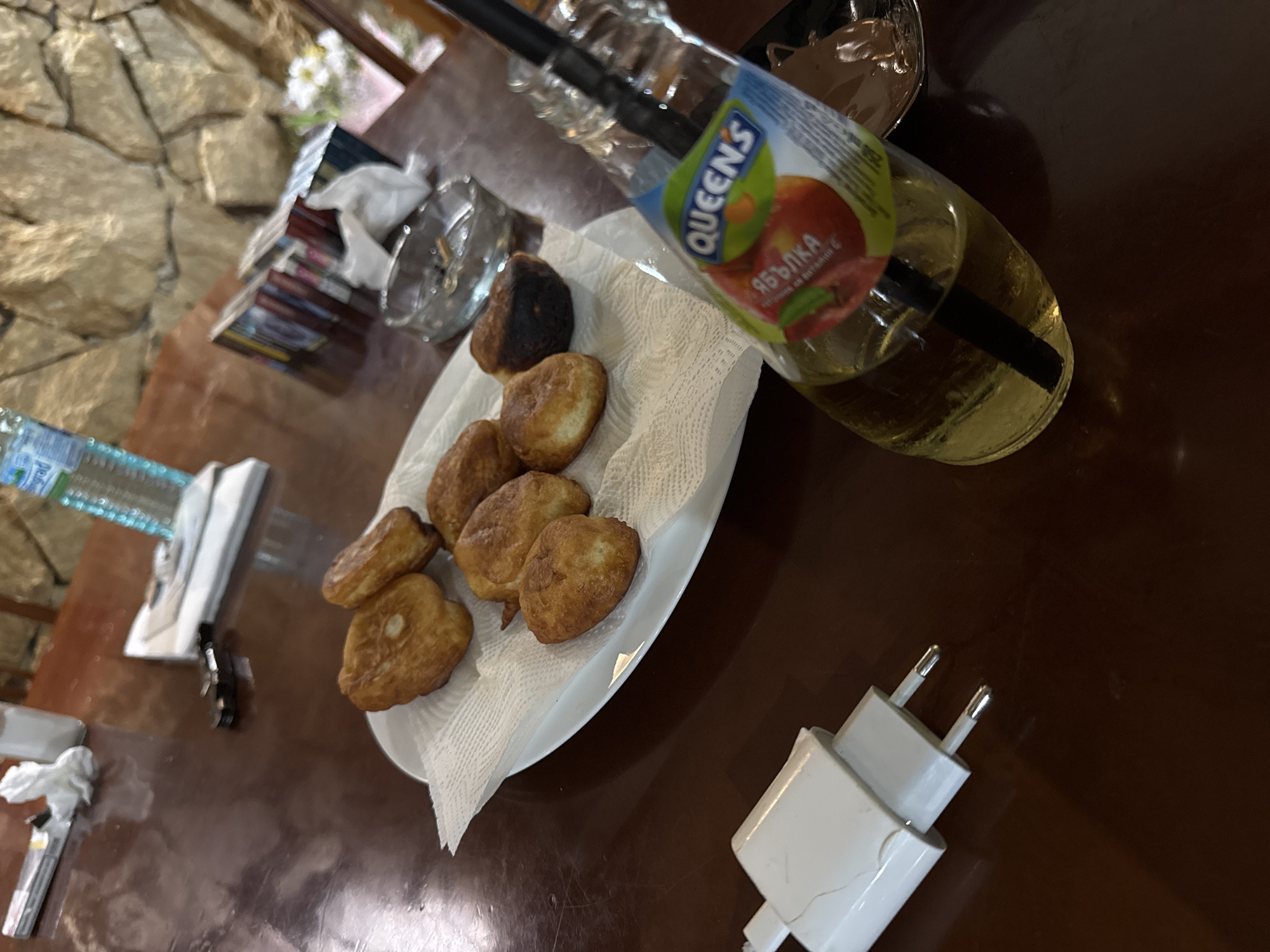 In Sredets, Bulgaria, an overhead shot captures a wooden table laden with various items. A white plate, resting on white napkins, displays a cluster of fried dough pastries. To the right, a glass bottle of Queens apple juice with a straw is positioned, its label featuring an apple graphic and Cyrillic text. Adjacent to the pastries is a clear glass ashtray containing cigarette butts. In the lower left corner, a white electrical adapter with two prongs is visible. The background reveals a stone wall and a selection of magazines. The lighting suggests an indoor setting, possibly a restaurant or cafe.