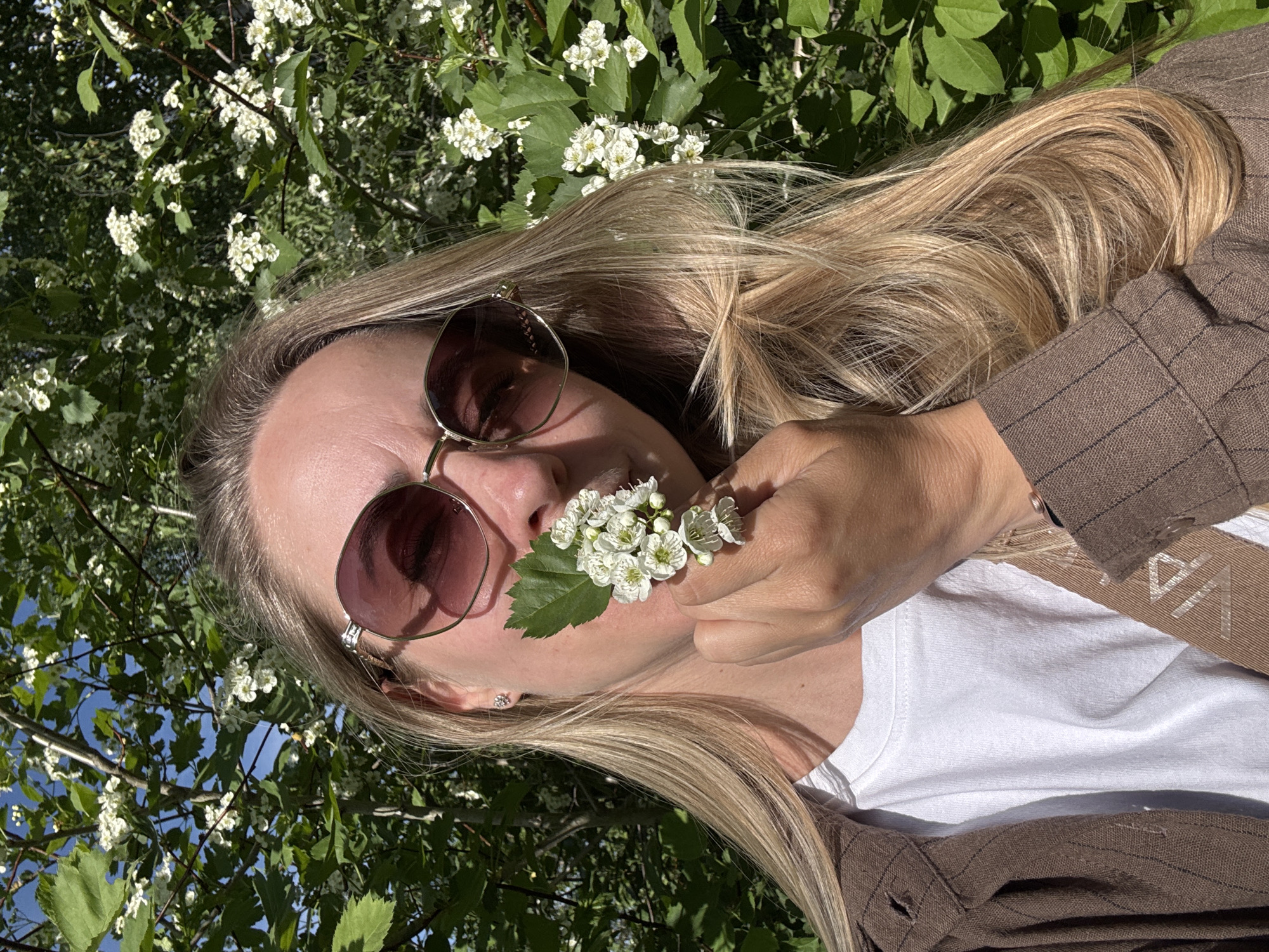A woman with blonde hair is holding a small cluster of white flowers near her face. She wears sunglasses with brown lenses and a brown striped jacket over a white t-shirt. The background is filled with green foliage and white flowers, with glimpses of blue sky visible through the leaves. The image appears to be taken in an outdoor, natural setting. While it is not possible to definitively confirm the location, the information provided suggests it could be in or around Chaikovsky, Russia.