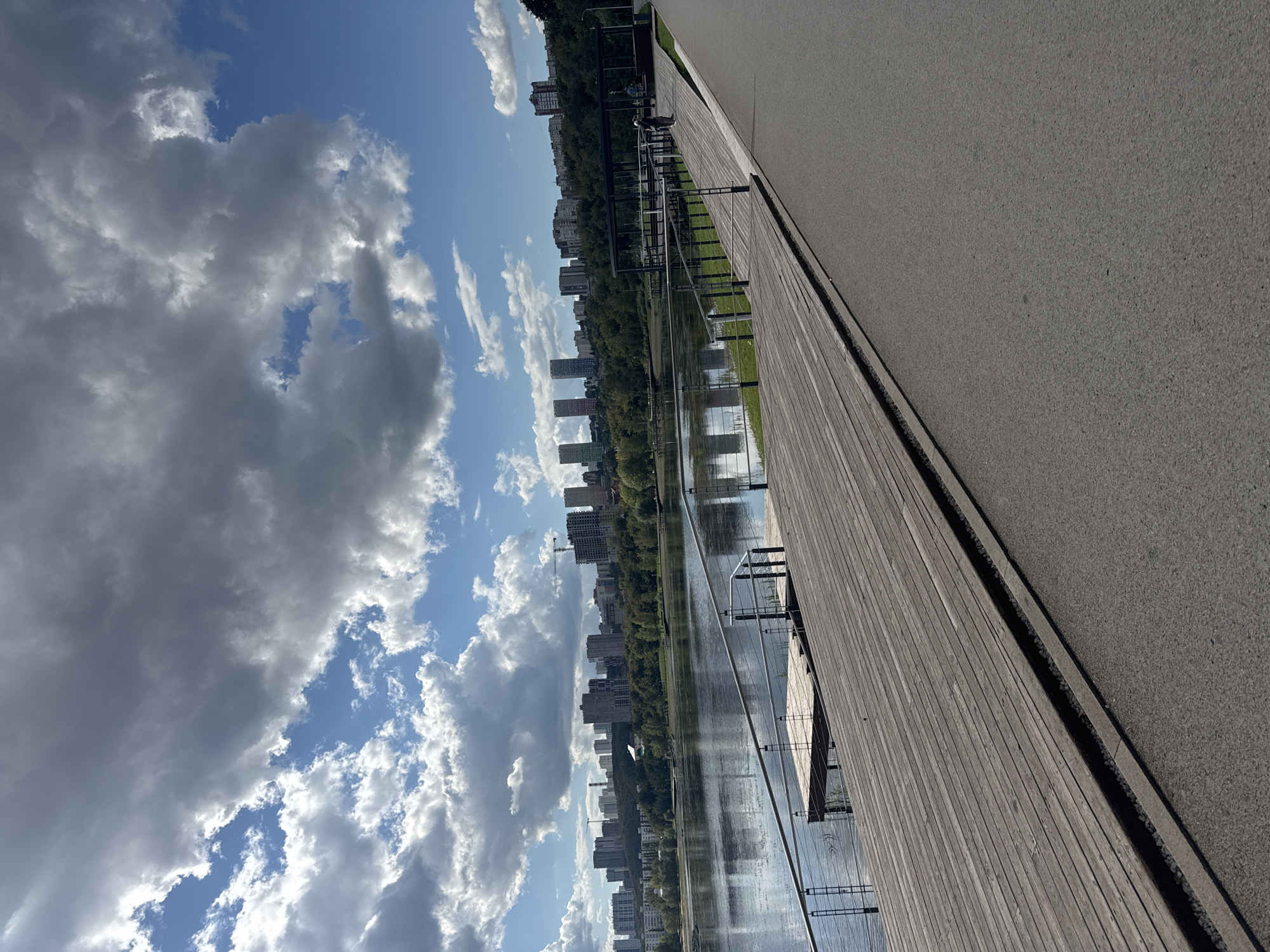 The image shows a waterfront promenade in Ufa, Russia, likely in the area of the National Museum of the Republic of Bashkortostan, overlooking a body of water, possibly a reservoir or a river. In the foreground, a wooden pier or boardwalk extends into the water. To the right of the boardwalk is an asphalt pathway, likely for walking or cycling. In the background, a cityscape with numerous residential and office buildings is visible, interspersed with green trees. The sky is partially cloudy with a mix of blue sky and dark, dramatic clouds. Two individuals are visible on the pathway in the middle ground, one appearing to be walking or standing near a covered structure. The overall scene suggests a public recreational area within an urban environment.
