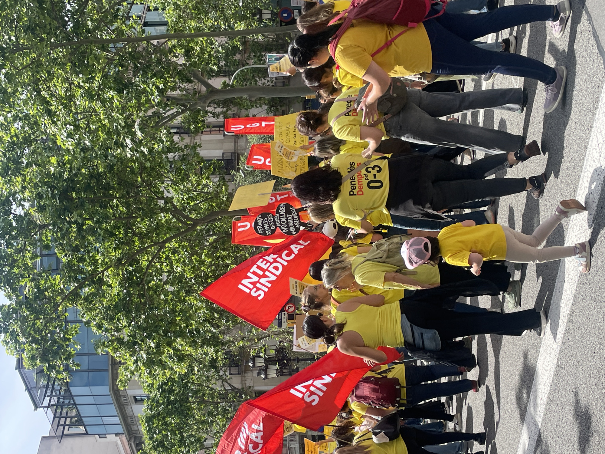 A medium-shot shows a demonstration in Barcelona, Spain, with several people marching on a street with buildings and trees. Most of the demonstrators are wearing yellow t-shirts. Some are holding red flags that read "Intersindical." Others hold yellow placards. One placard features the phrase "Penedes dempes pel 0-3." The demonstration takes place on a city street with crosswalk markings. Tall trees with thick foliage are visible on the left side of the frame. A modern building with glass windows is partially visible behind the trees. The scene appears to be brightly lit, suggesting a sunny day.