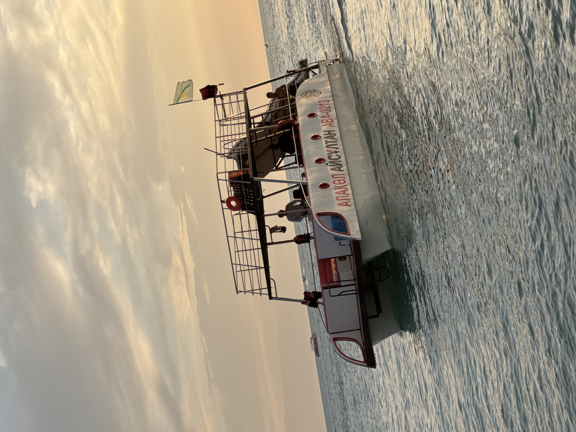 A catamaran boat, identified by the Cyrillic text "АЛАКӨЛ АЙСҰЛТАН АВА-0213" on its side, cruises through calm, rippling water. The boat features a white hull with red accents and a two-tiered passenger deck with metal railings. Several individuals are visible on the upper deck, appearing to be passengers or crew. One person stands at the helm. A flag, possibly the flag of Kazakhstan, flies from a pole on the boat's roof, near a life preserver. In the background, other smaller boats are visible on the water, and a hazy, mountainous horizon can be seen against a soft, pale sky with scattered clouds, illuminated by the warm glow of sunset. The overall scene suggests a leisurely excursion on the water, likely at Alakol Lake in the Zharbulak region of Kazakhstan, given the boat's inscription and the landscape.