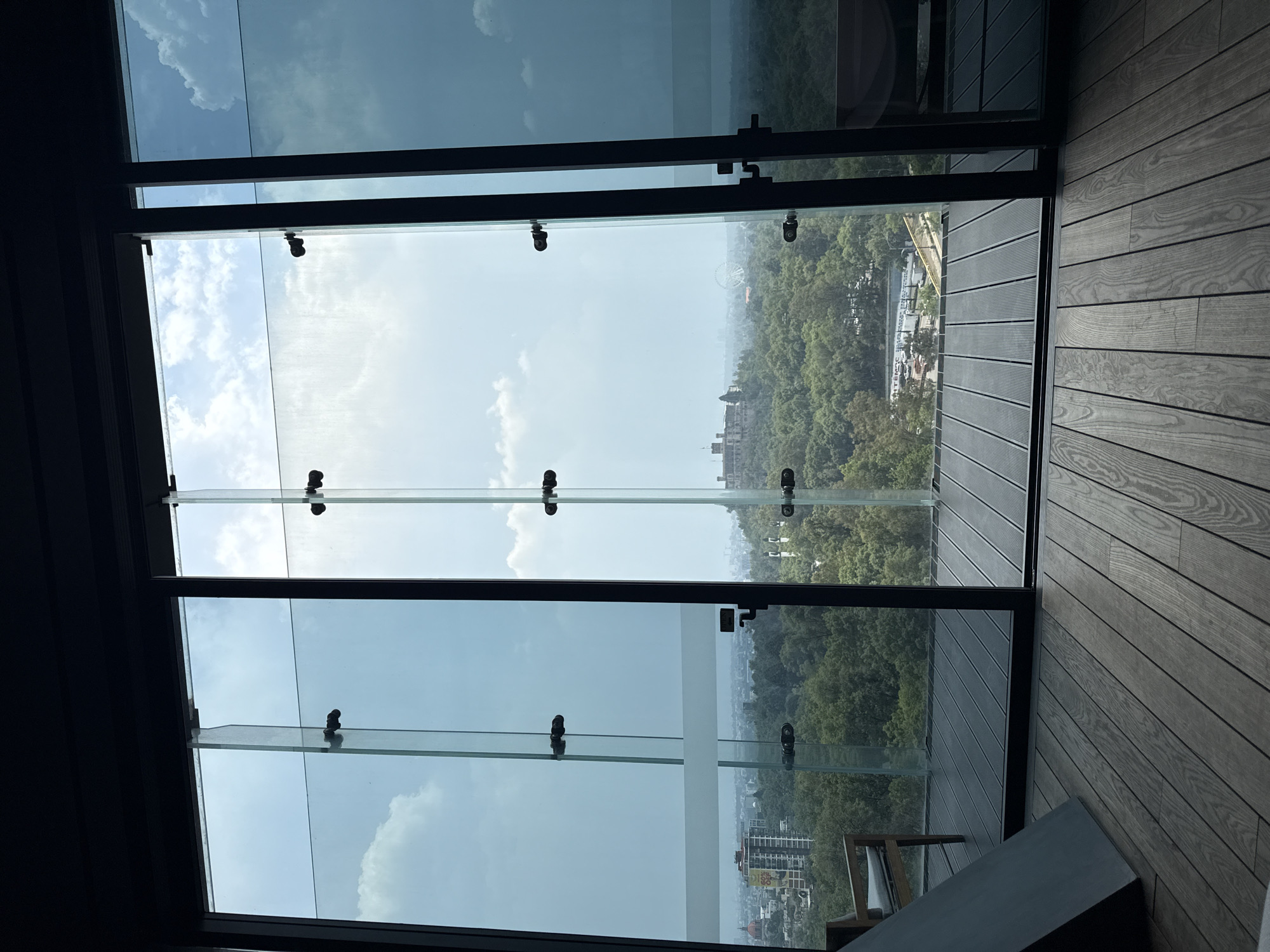 The image presents an elevated view of Mexico City, Mexico, through a window comprised of multiple glass panes held together with metal fittings. The window opens onto an outdoor area with grey wooden decking.
Beyond the glass, a cityscape of Mexico City is visible. The foreground features dense green trees that transition into a more urban landscape with buildings. The sky is partially cloudy with bright sections.