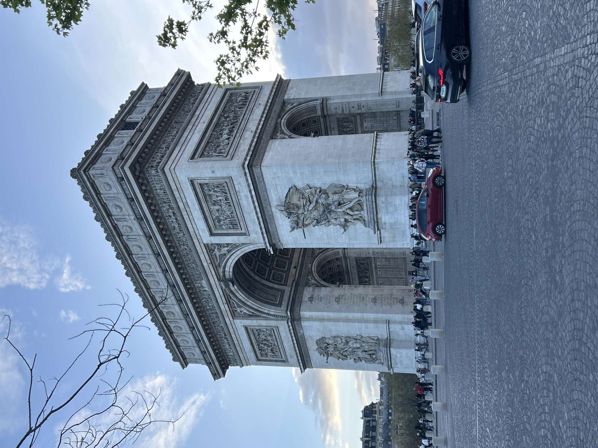 The image is a photograph of the Arc de Triomphe, located in Paris, France. The Arc is a large, gray stone structure with intricate carvings. There is a cobblestone paved road in front of the Arc, and a line of people are waiting behind a barrier. Several cars are parked on the road. There are trees visible in the background and foreground. The sky is blue with white clouds.