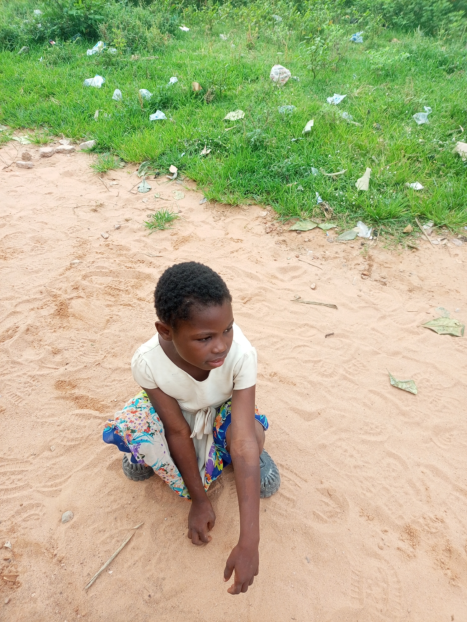 A young Black girl with short, dark, curly hair sits in a sandy area with green grass and scattered debris in the background. She is wearing a cream-colored dress and a colorful, patterned skirt. Her gaze is directed to the right, and her arms are resting on her legs. The ground is composed of light brown sand with visible footprints. The background features lush green grass littered with white and brown plastic trash.