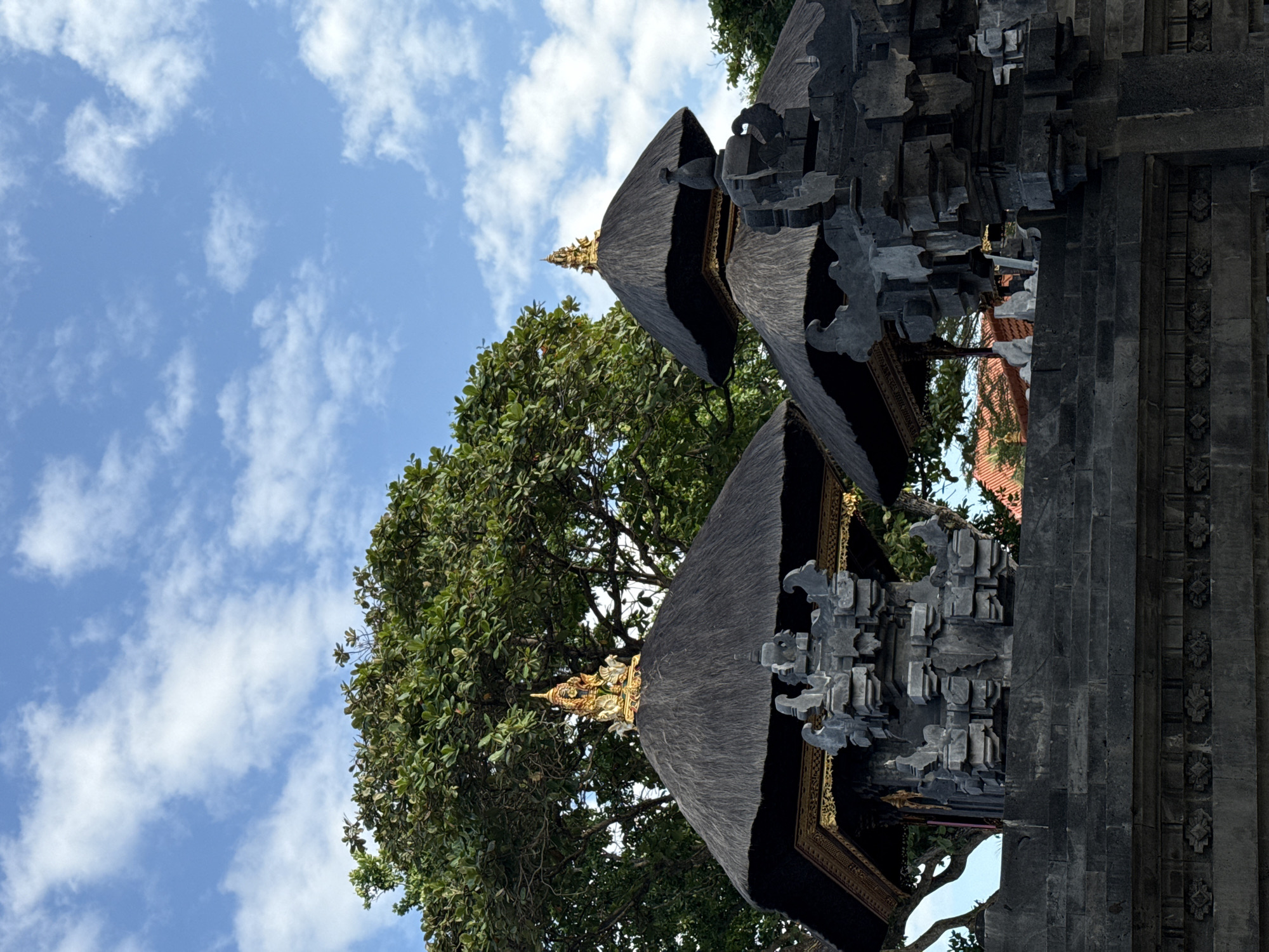 The image displays traditional Balinese temple architecture situated in Kuta Selatan, Indonesia. In the foreground and midground, dark grey volcanic stone structures with intricate carvings are visible, forming what appears to be part of a gate or decorative wall. Behind these, two distinct pavilions with dark, conical thatched roofs, likely made of ijuk fiber, stand prominent. The pavilion on the left features a broader thatched roof, surmounted by an elaborate golden deity figure on a golden base. The pavilion on the right has a taller, more pointed thatched roof, capped with a dark, unadorned finial. Both pavilions have dark structural bases beneath their roofs, accented with golden trim. Lush, dense green foliage from large trees provides a natural background, partially visible behind and between the structures. The upper portion of the image reveals a clear blue sky scattered with white cumulus clouds. No people or animals are visible.