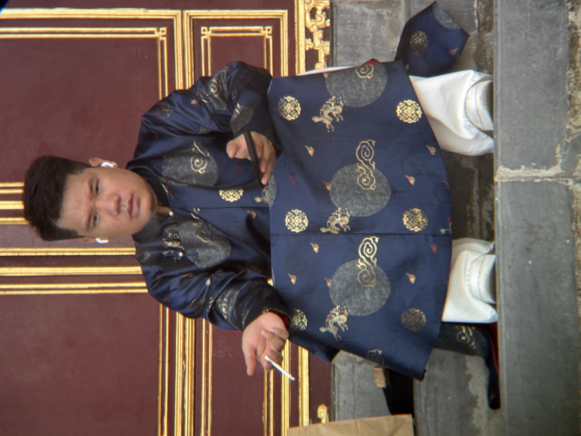 A person wearing a dark blue traditional garment with gold embroidery and white pants sits on a stone step in front of a maroon and gold ornate door, holding a cigarette in their right hand and a dark rectangular object, possibly a phone, in their left. The person has short dark hair and is wearing white earbuds. The scene is likely in Phú Xuân Ward, Vietnam, given the architectural style.