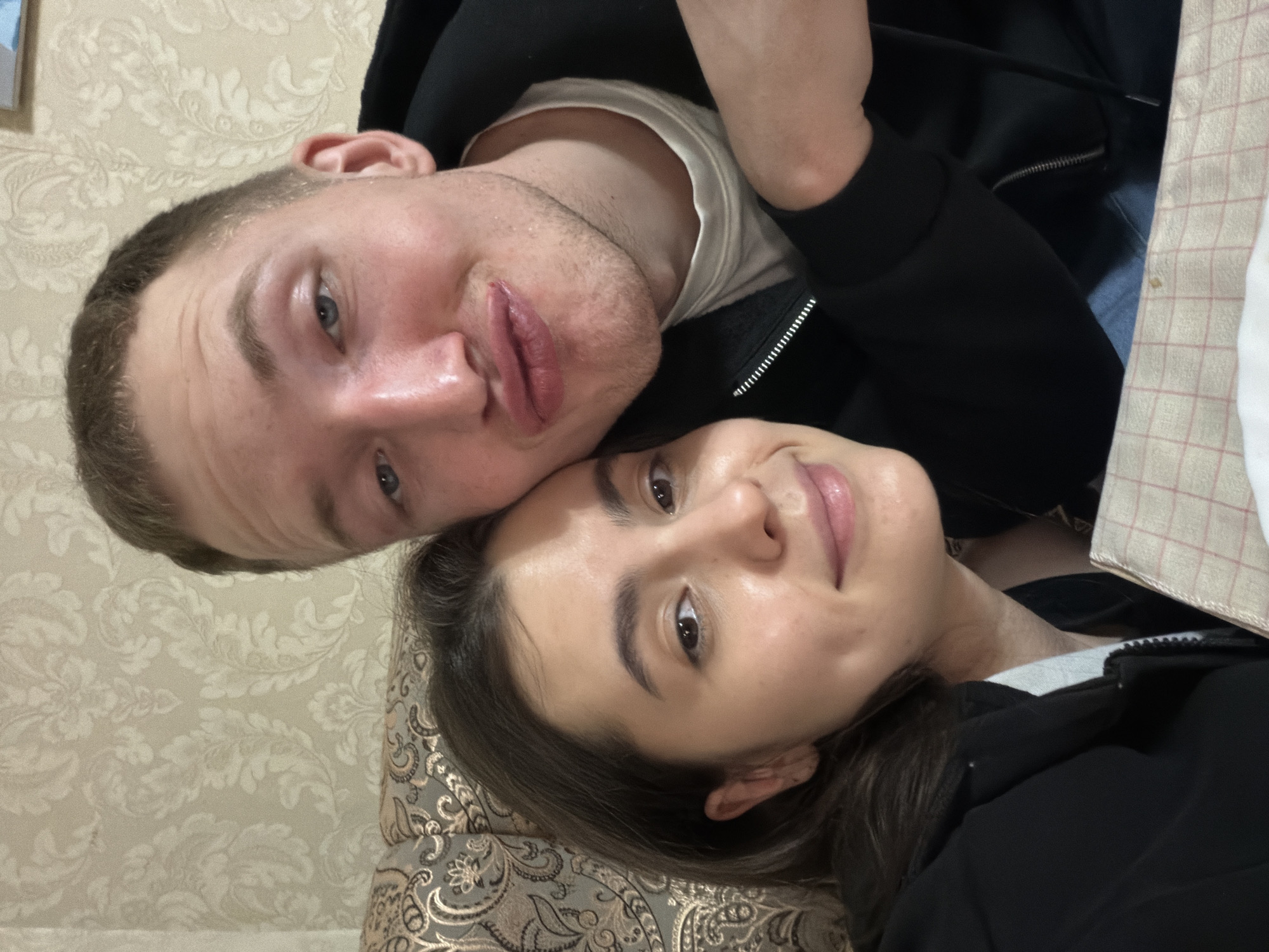 A close-up, rotated selfie depicts a man and a woman posing cheek-to-cheek. The man, located at the top left of the frame, has short, light brown hair, fair skin, and blue eyes. He wears a black hoodie over a white t-shirt. His expression is somewhat neutral with slightly pursed lips. The woman, situated at the bottom right, has dark brown hair, fair skin, and brown eyes. She also wears a black hoodie over a lighter-colored garment. She offers a subtle smile. The background consists of a light-colored wall with a floral pattern. The location is Chaikovsky, Russia. A decorative patterned pillow is partially visible beneath the woman.