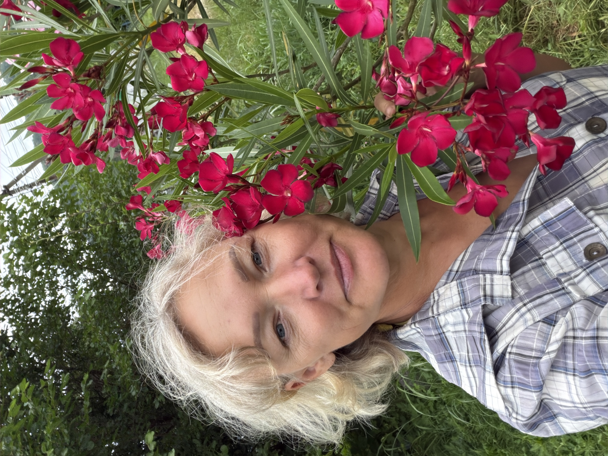 The image shows a middle-aged Caucasian woman with blonde, wavy hair and blue eyes. She is wearing a blue and white plaid shirt and is standing outdoors, near a bush with vibrant pink flowers. The background consists of lush green foliage and grass, suggesting a natural or garden setting. The woman is looking towards the camera with a slight smile. There is no specific indication of a location within the city of Shepsi, Russia.
