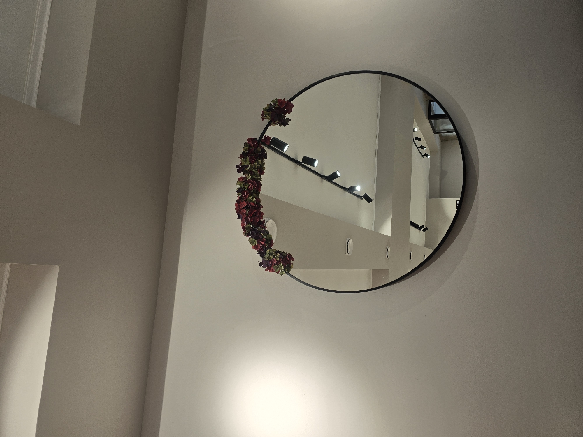 An interior shot shows a round mirror with a black frame, decorated with a garland of red and green artificial flowers at the top. The mirror reflects a ceiling with recessed lighting. The wall behind the mirror is a neutral, light color, with a large area of illumination on the left side, possibly from a window or a strong light source. The image does not provide any information that would suggest the location is in Ulan-Ude, Russia.
