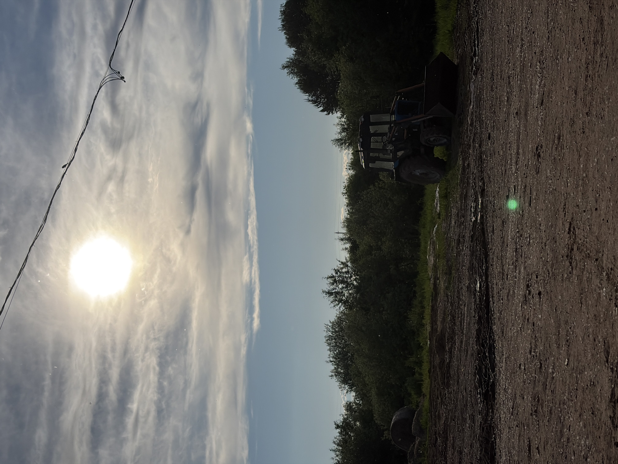 The image captures an outdoor, rural, or semi-rural scene, likely in Chumna, Russia, oriented with the sky on the left and the ground on the right. When viewed correctly, the upper portion of the frame features a bright, intense sun high in the sky, casting a strong glare. The sky is a mix of clear blue and wispy, scattered clouds.
A power line with small attachments or insulators stretches diagonally across the upper left quadrant of the sky. Below the horizon line formed by the sky, a dense line of dark green trees and foliage extends across the middle of the image. On the right side, a dirt or gravel road occupies the foreground and midground, with some green grass and vegetation bordering it. A dark-colored tractor or heavy machinery, possibly a front-end loader, is partially visible behind the road and amidst the trees on the left side. A prominent bright green lens flare is visible on the dirt road, indicating the direct sunlight.
The scene depicts a bright, clear day, suggesting morning or afternoon given the sun's position. No people or specific activities are visible. There is no visible text in the image.