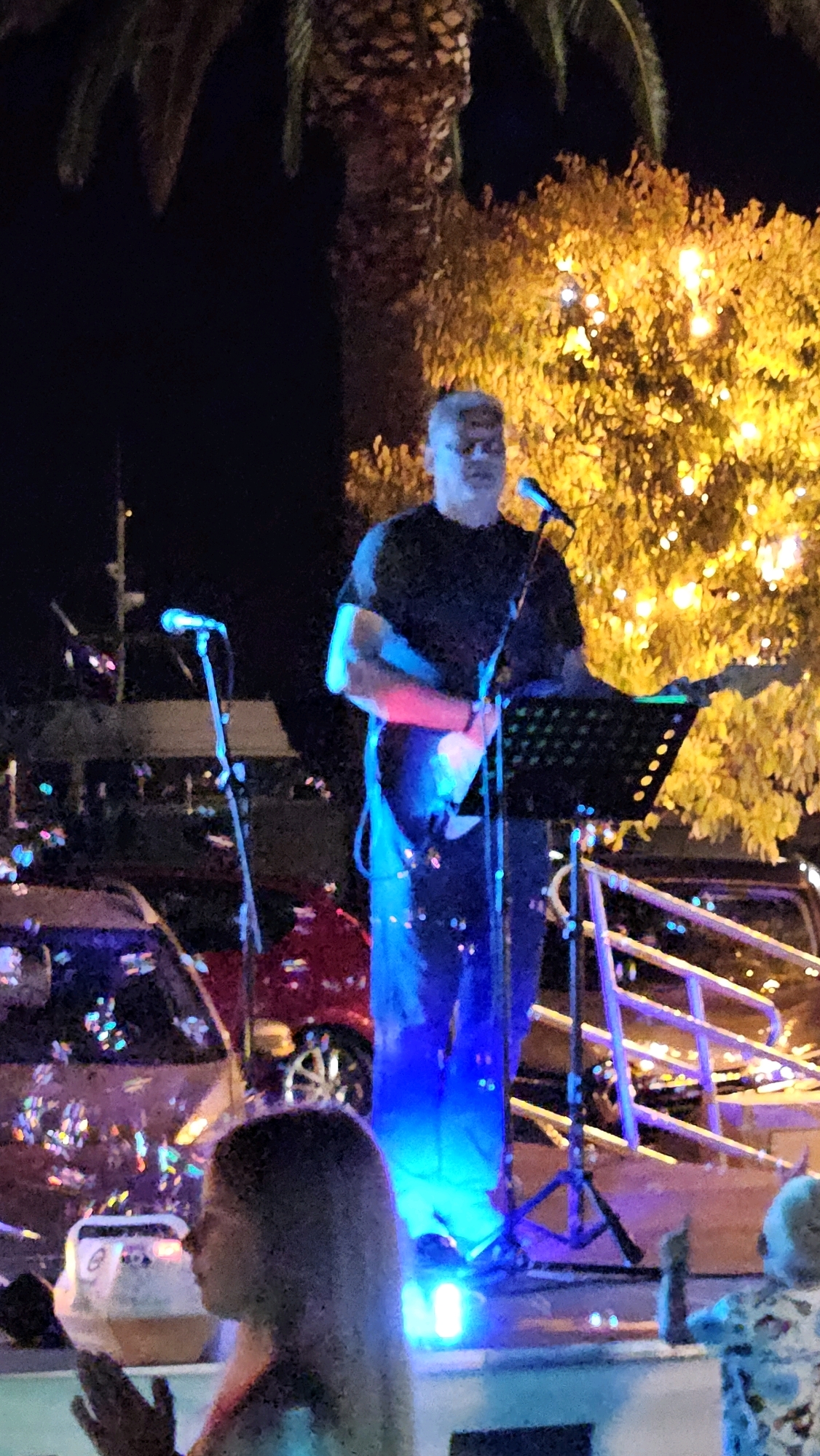 The image depicts a nighttime scene at an outdoor venue, likely a concert or performance, in Općina Tučepi, Croatia. A male musician, appearing to be in his late 50s or early 60s with graying hair, is playing an electric guitar on a stage illuminated by blue and pink stage lights. He is wearing a dark t-shirt and dark pants. A microphone stands are positioned on the stage. In the background, a tree adorned with bright yellow lights is visible, along with a dark sky. To the left of the stage, a red car is partially visible, and further back, a railing with lights can be seen. In the foreground, the backs of two people's heads are visible, one with long hair, suggesting an audience watching the performance. The overall atmosphere appears festive and lively, typical of a summer evening event in a tourist destination.