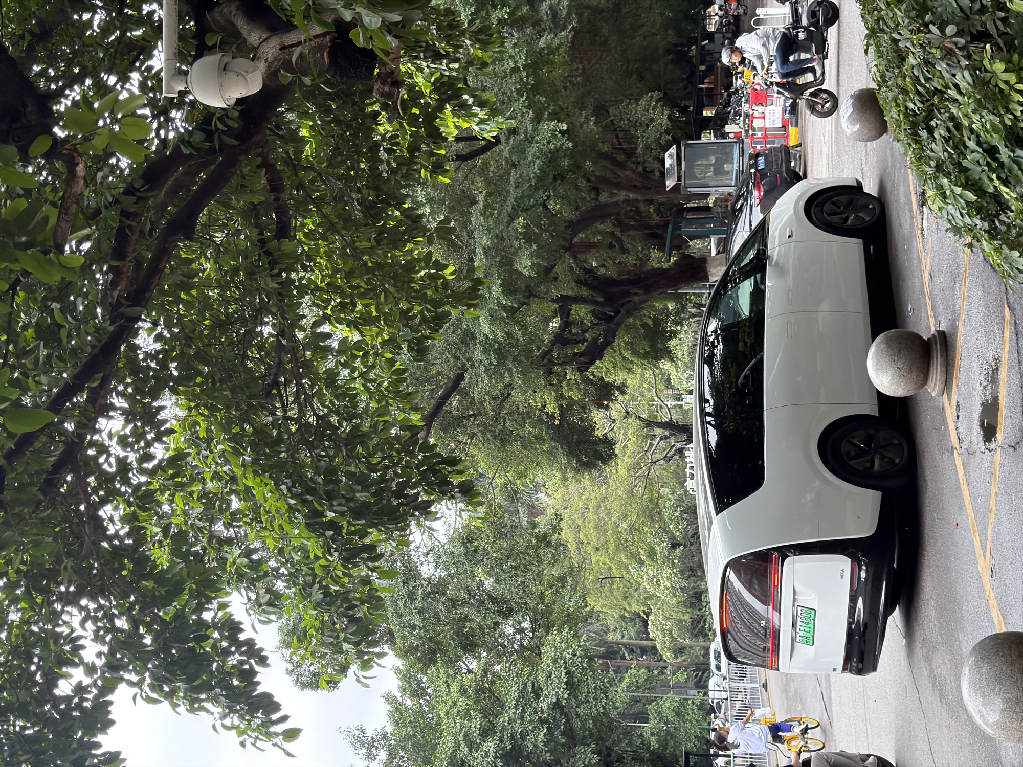The image shows a street scene in what appears to be the Baiyun District in China, indicated by the license plate "粤A" from Guangzhou. A white car is parked along the side of the road near a row of stone bollards. The environment is lush with trees. In the background, there are parked scooters and a person is seen riding a scooter. A security camera is affixed to a tree branch.